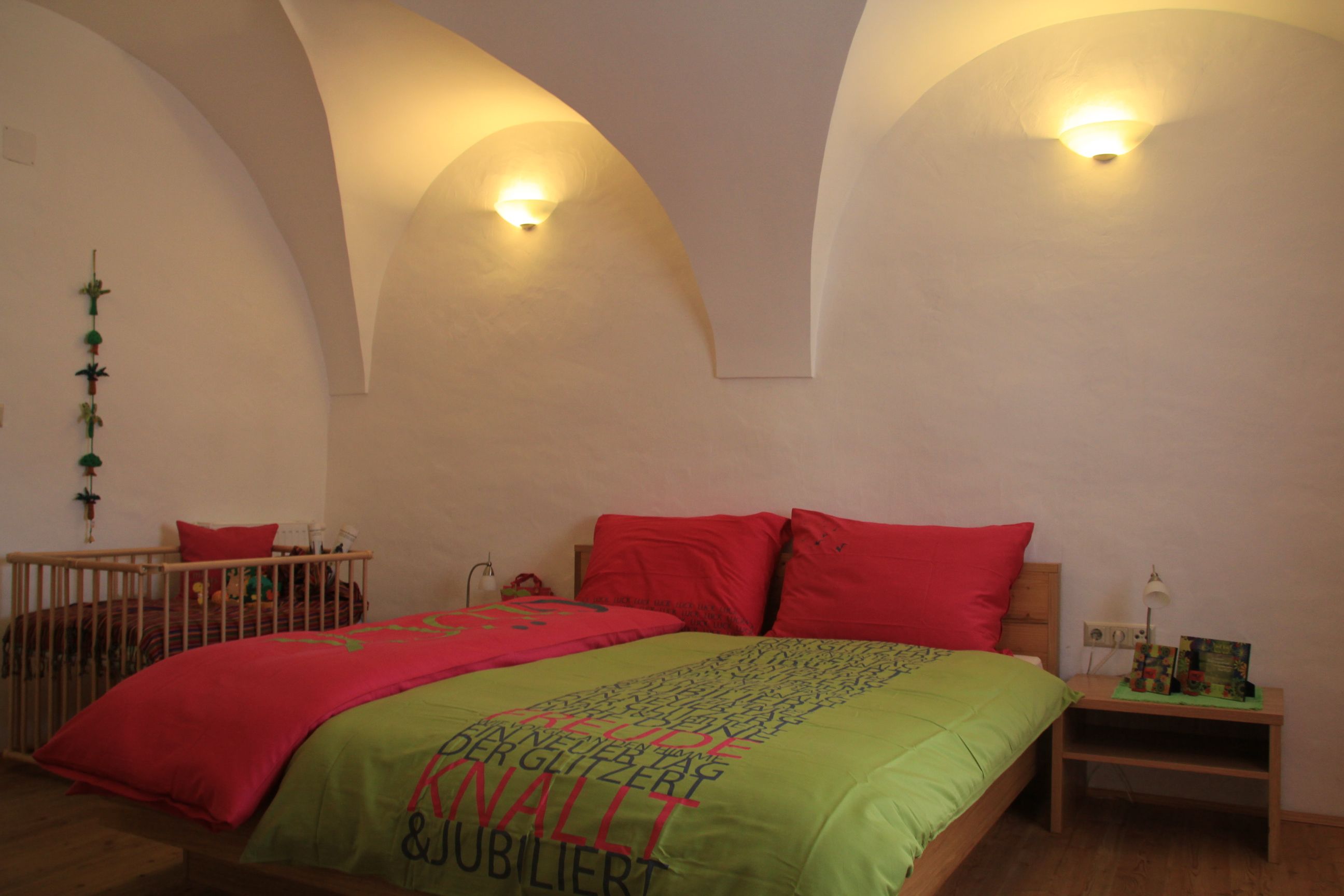 Útulná izba pre hostí s manželskou posteľou, farebnými perinami a detskou postieľkou v pozadí. Steny sú biele a klenuté.