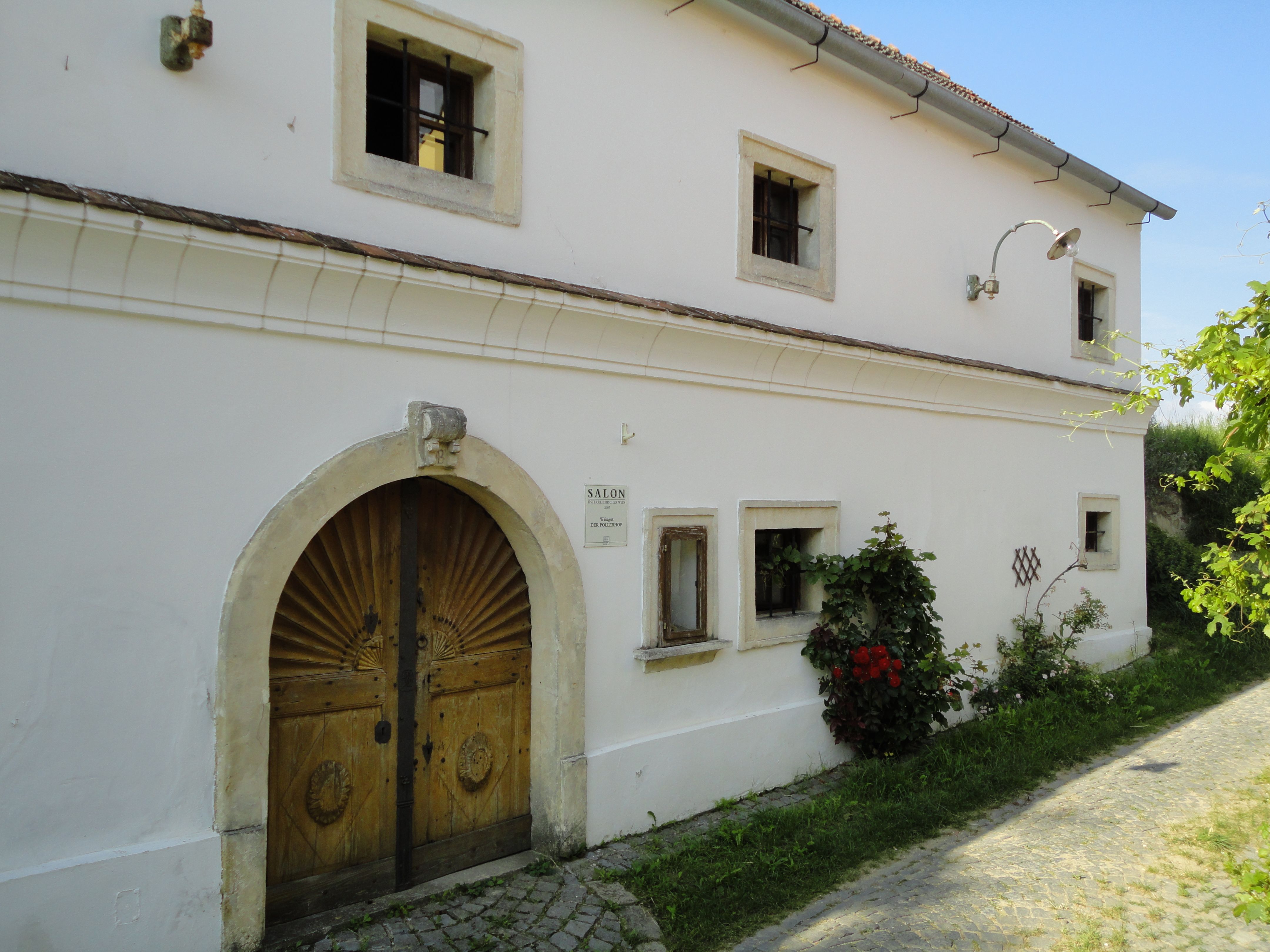 Historická budova s drevenými dverami a malými oknami, obklopená rastlinami.