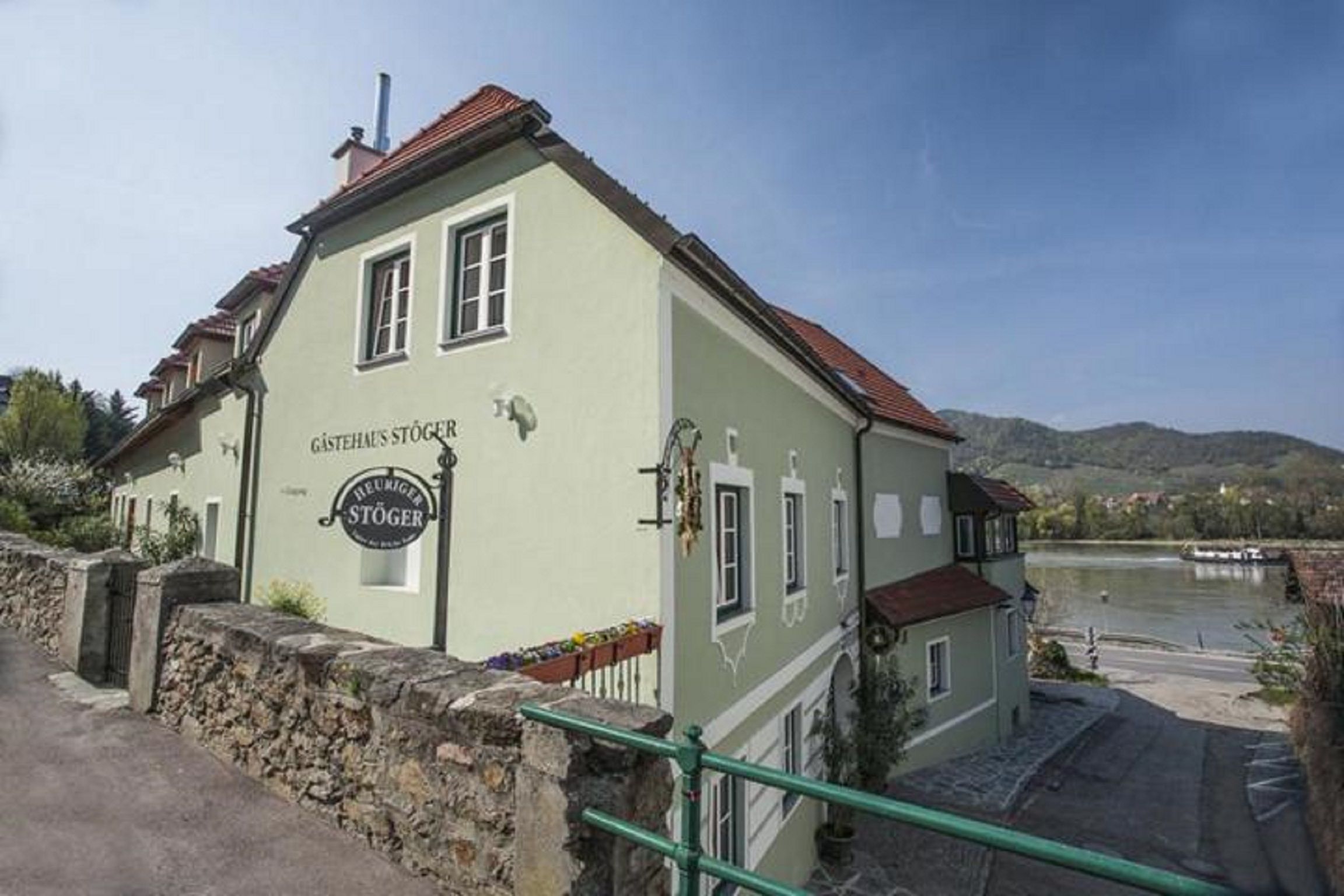 Zelený penzión s označením "Gästehaus Stöger" a "Viecha Stöger" na brehu rieky.
