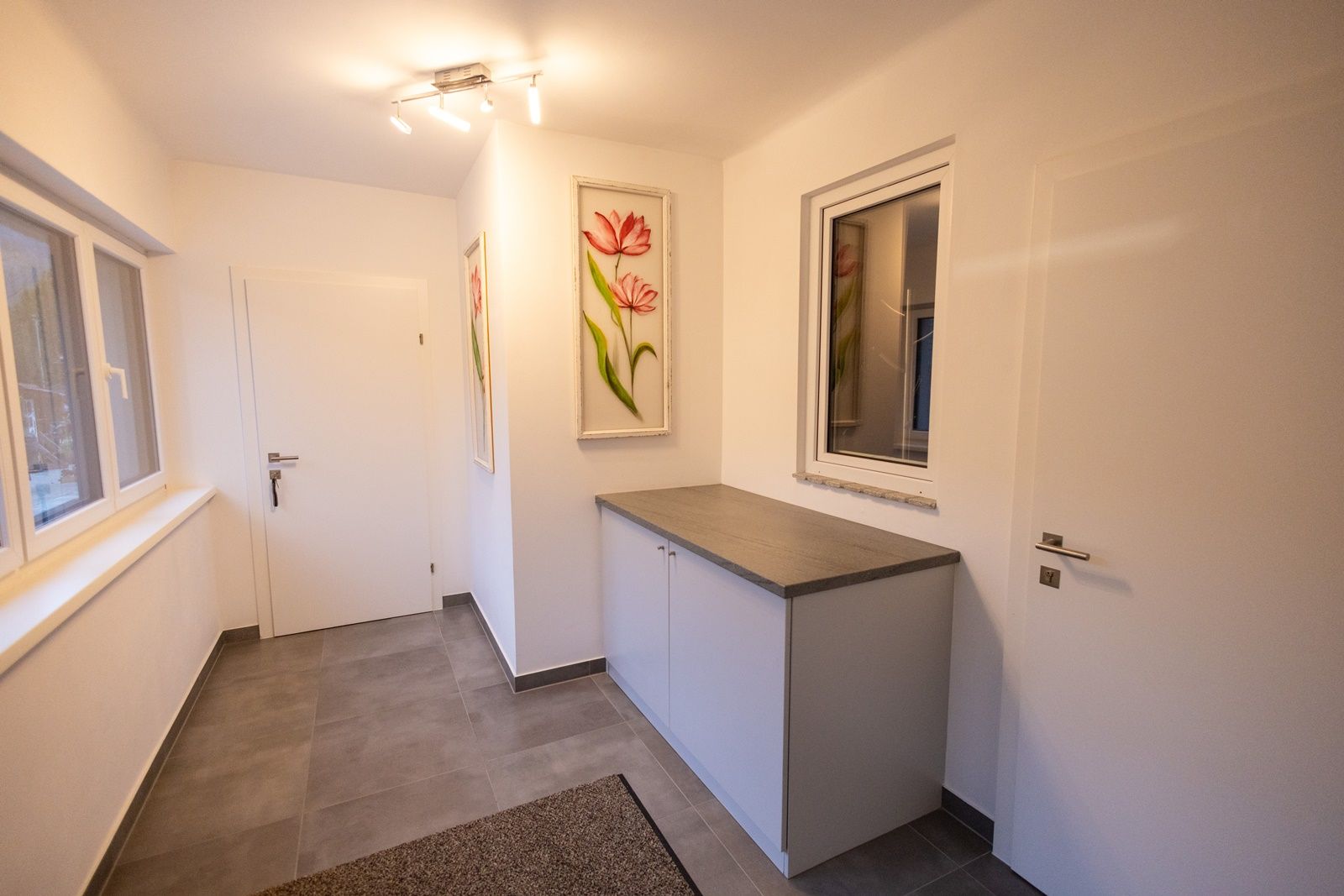 Moderná vstupná hala s bielymi stenami, sivou podlahou, skriňou a obrázkami kvetov na stene.