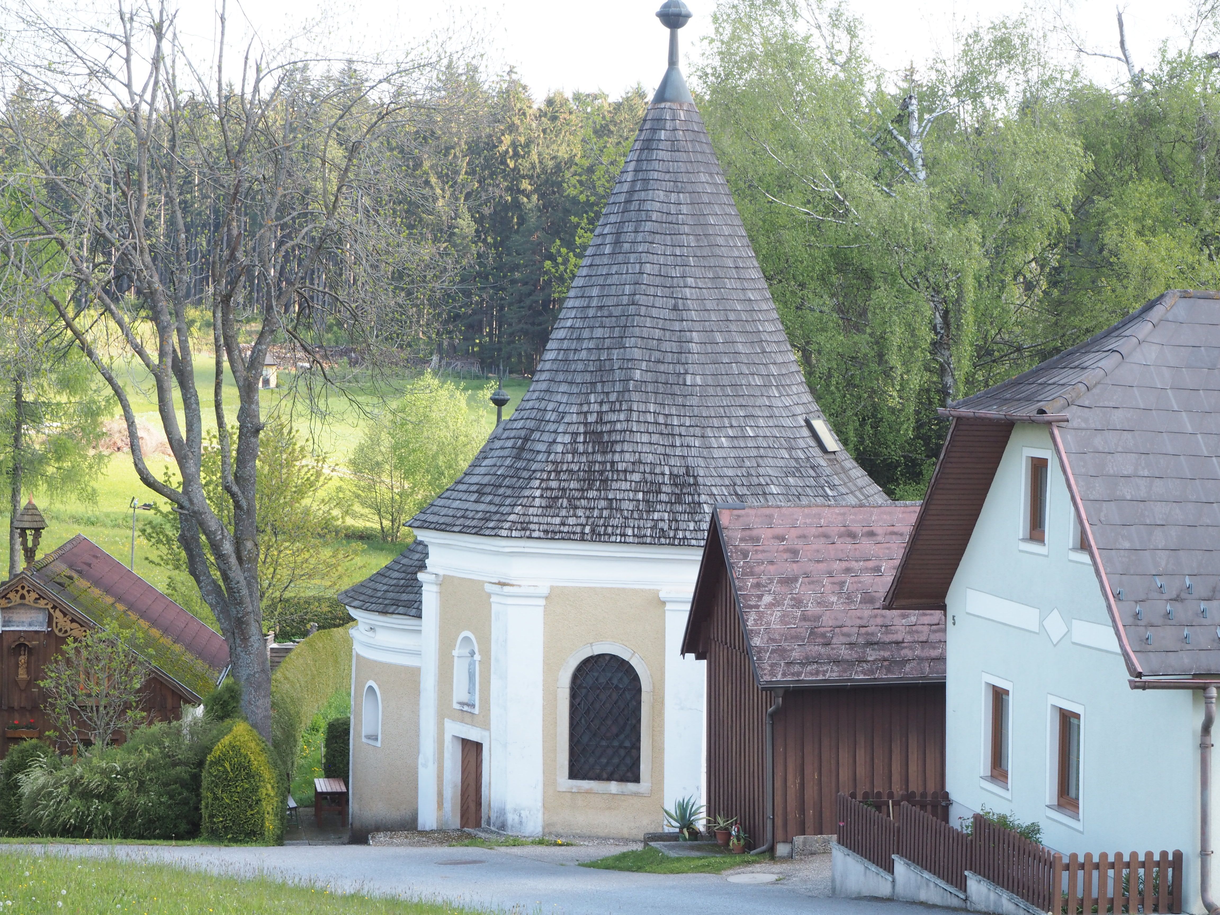 Malá kaplnka so špicatou strechou, obklopená stromami a domami.