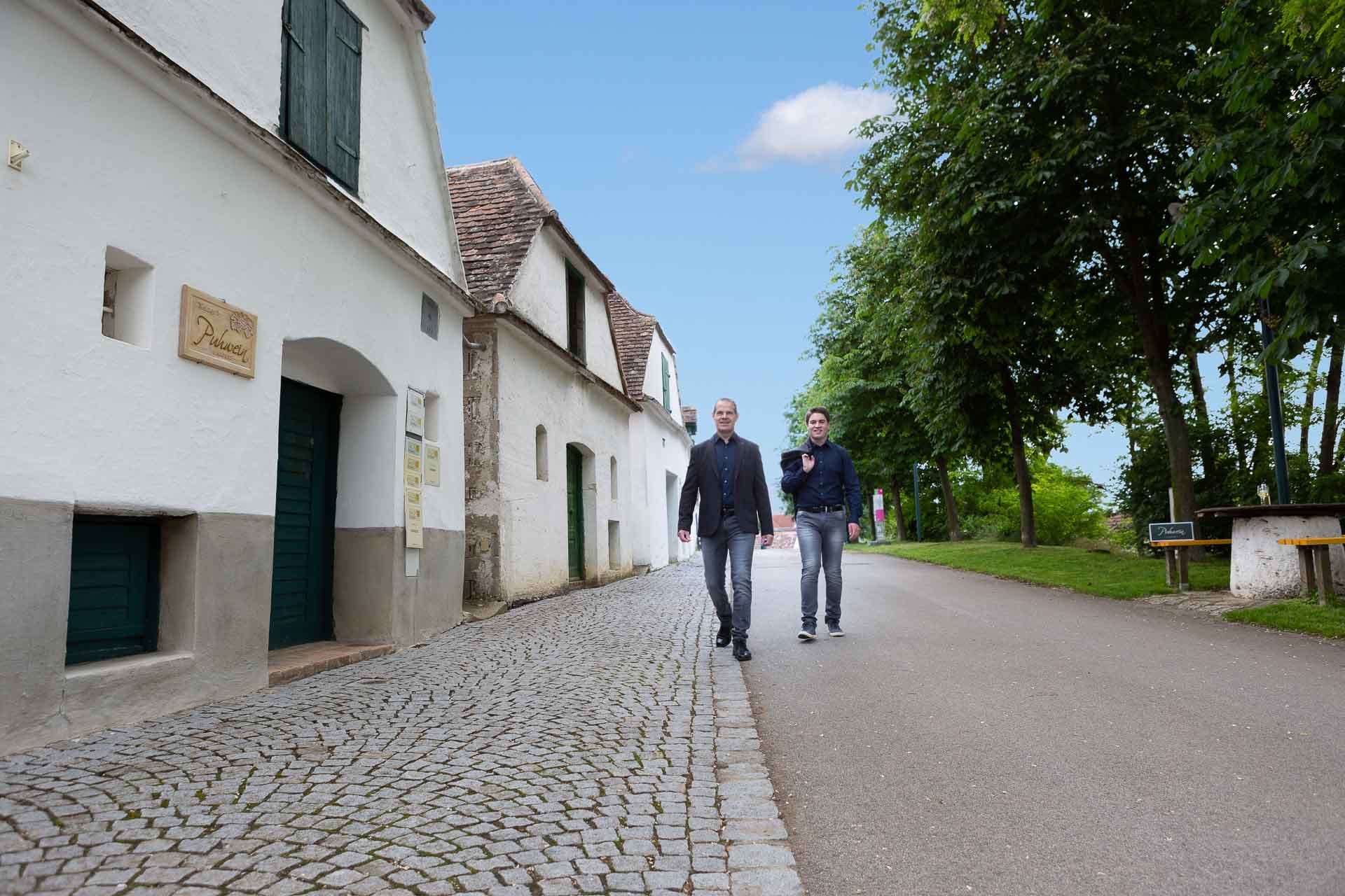 Dvaja muži kráčajú po dláždenej ulici okolo bielych budov a stromov.