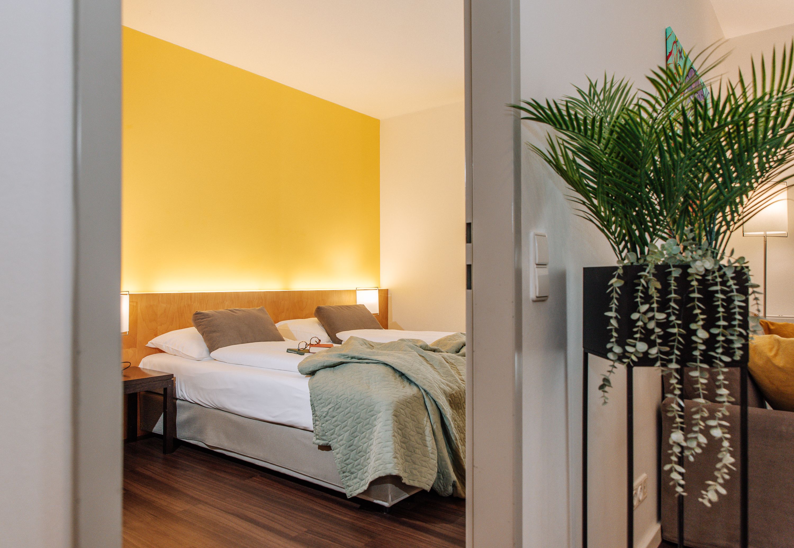 Hotelová izba so žltou stenou, manželskou posteľou a rastlinou v popredí.