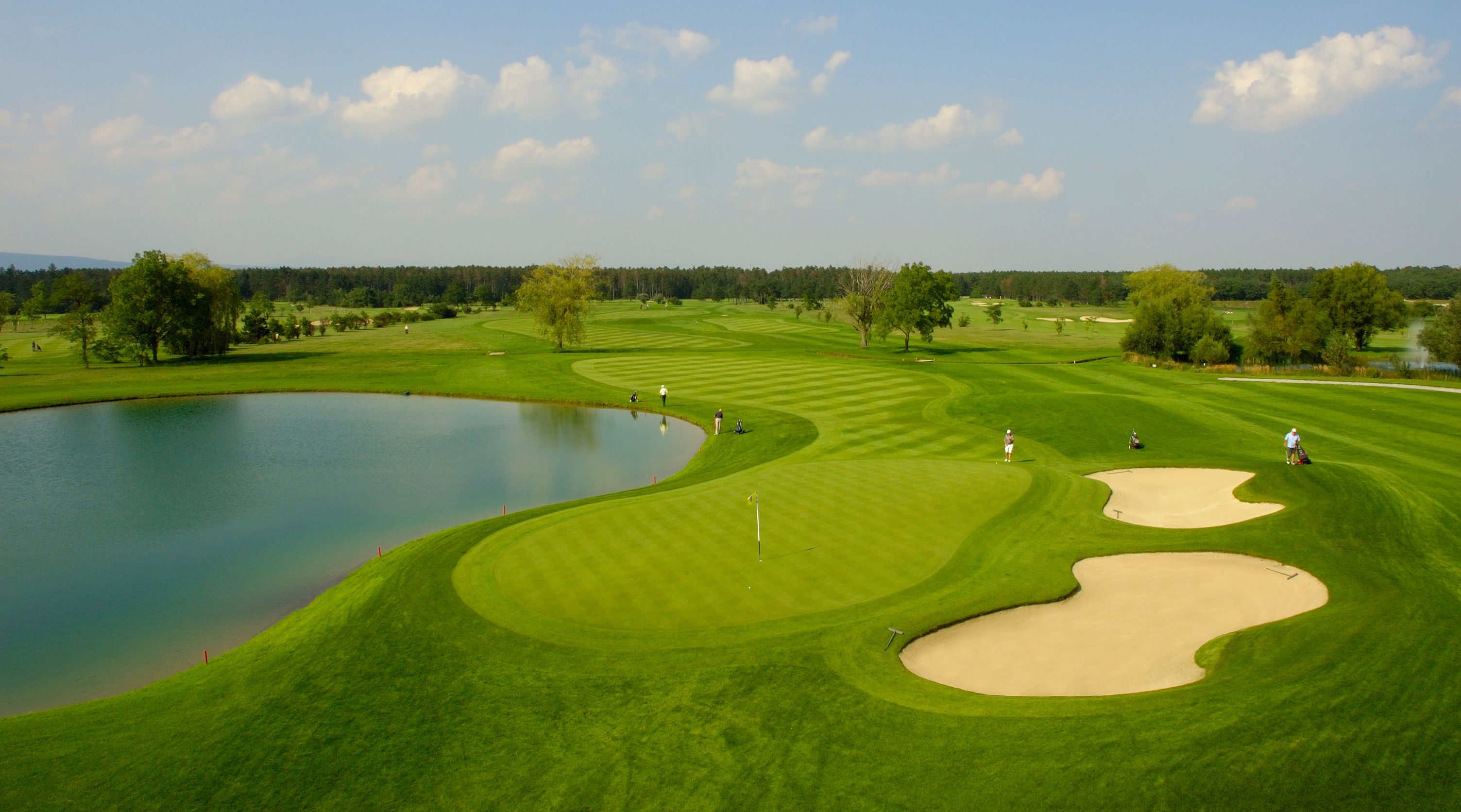 Golfové ihrisko s rybníkom, pieskovými bunkrami a hráčmi.