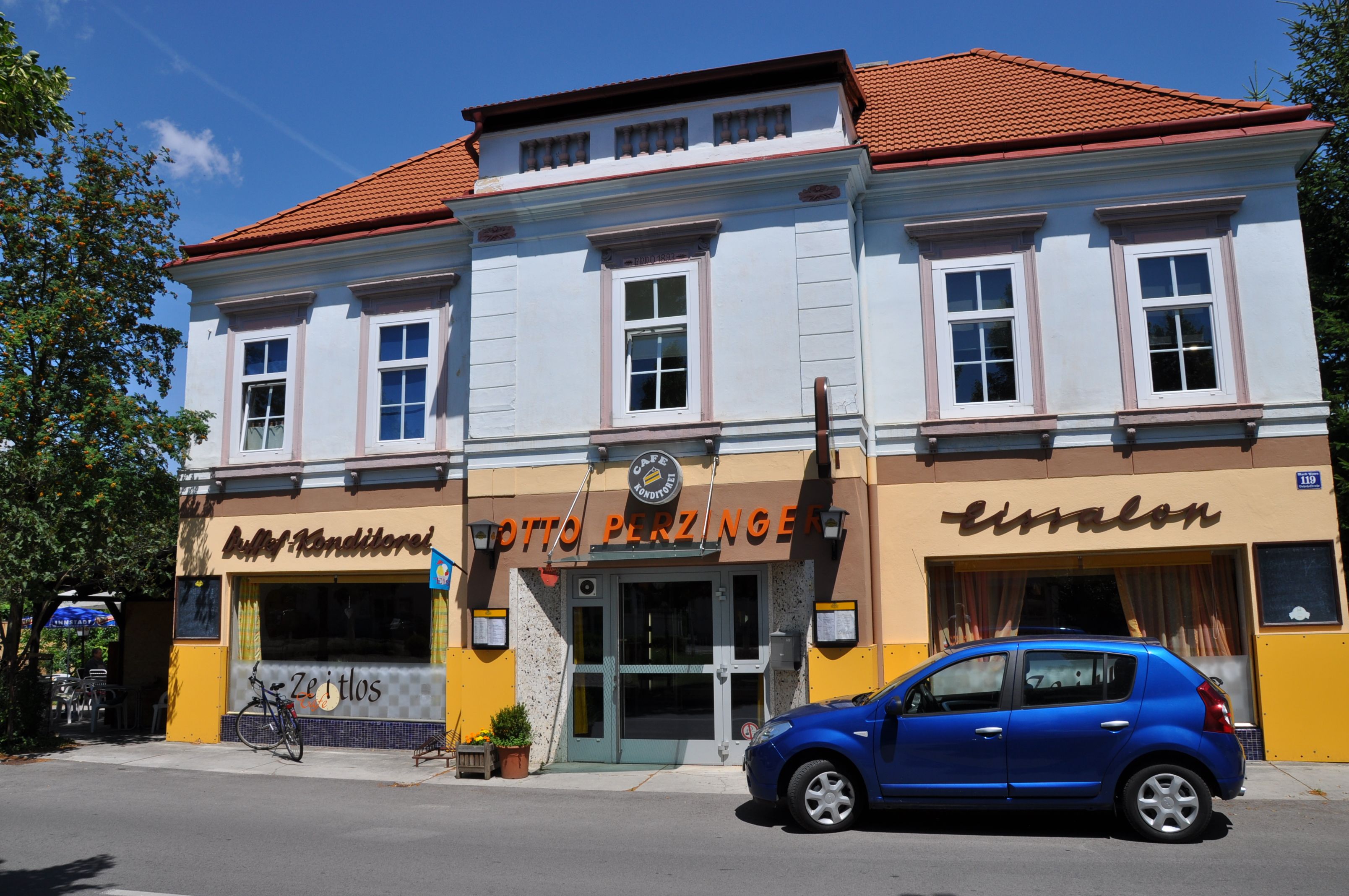 Pohľad zvonku na kaviareň s modrou fasádou a červenou strechou, pred ktorou parkuje modré auto.