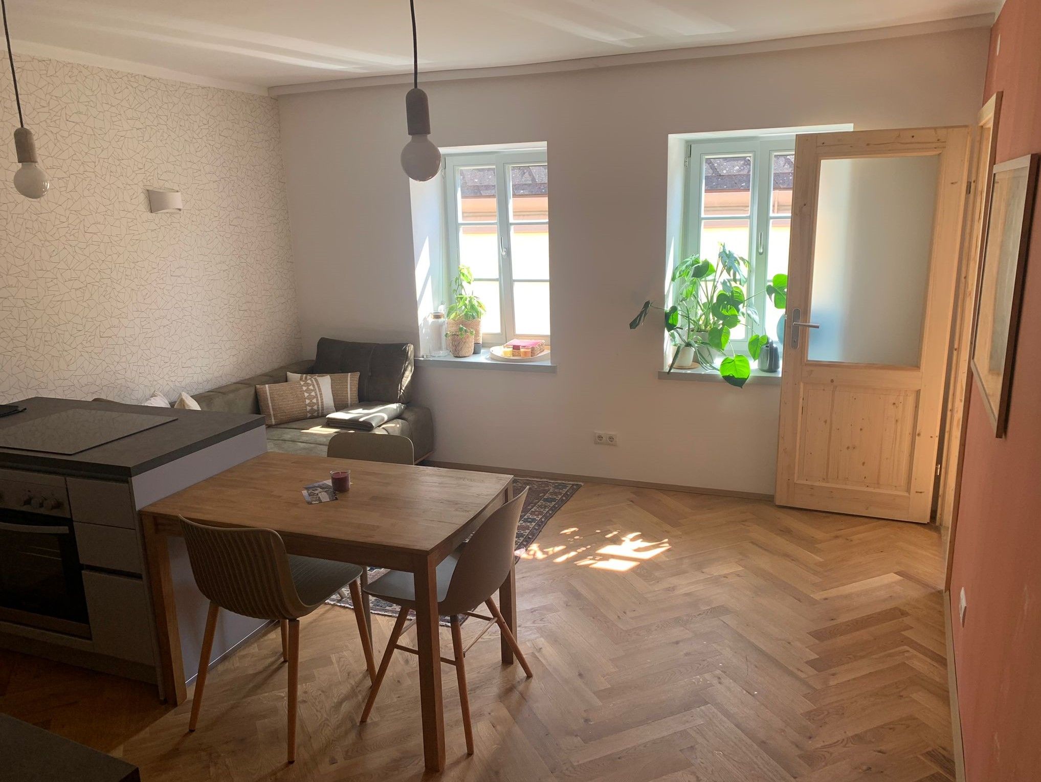 Svetlá obývacia izba s drevenou podlahou, jedálenským stolom, pohovkou a rastlinami pri okne.