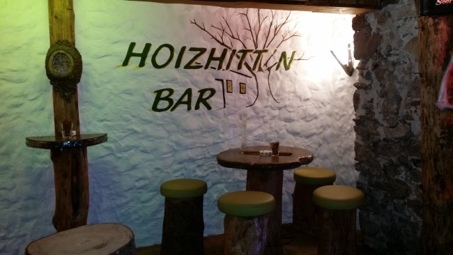 Rustik&aacute;lny bar s dreven&yacute;m n&aacute;bytkom a n&aacute;pisom "Hoizhitt'n Bar" na stene.