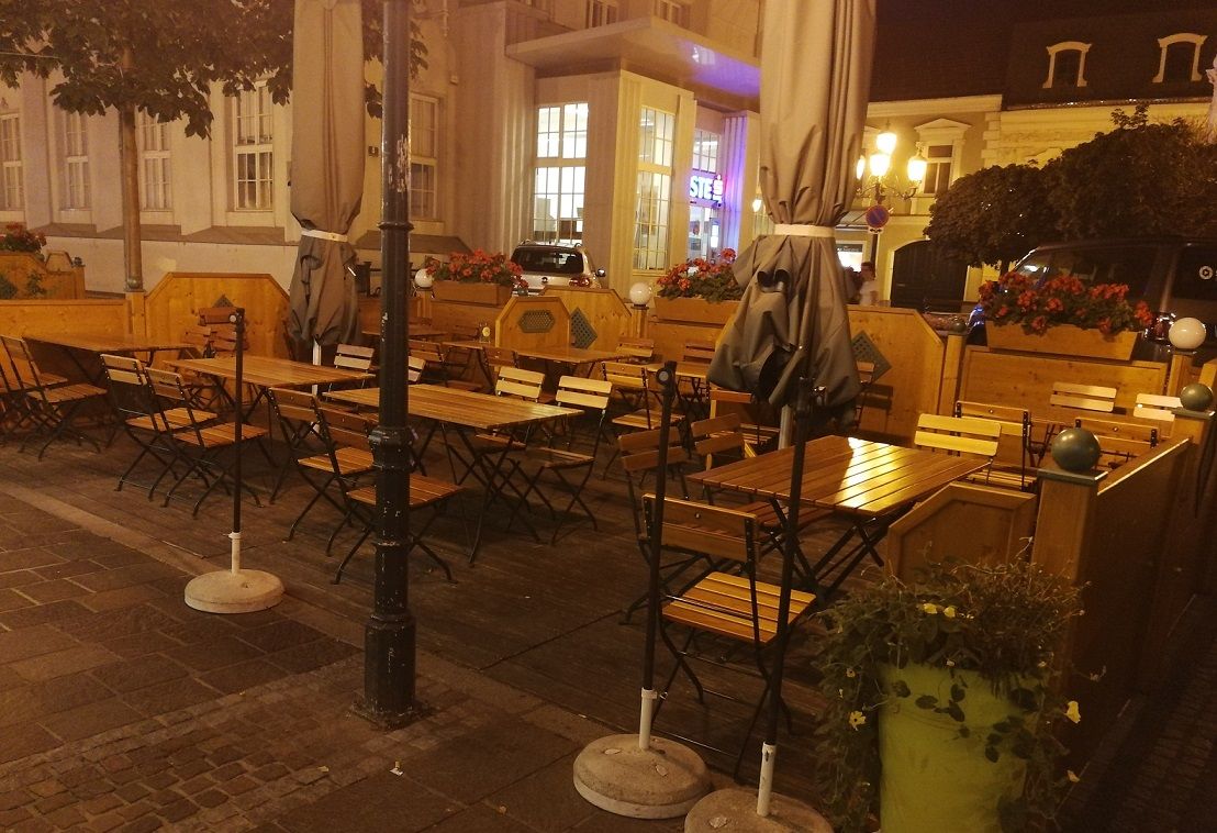 Vonkajší priestor reštaurácie s prázdnymi drevenými stolmi a stoličkami, v noci obklopený rastlinami a lampášmi.