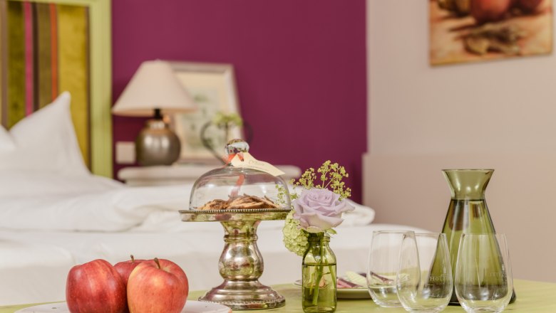 &Scaron;t&yacute;lovo zariaden&aacute; miestnosť so stolom, na ktorom s&uacute; umiestnen&eacute; jablk&aacute;, su&scaron;ienky pod sklenen&yacute;m zvončekom, v&aacute;za s kvetom a poh&aacute;re.