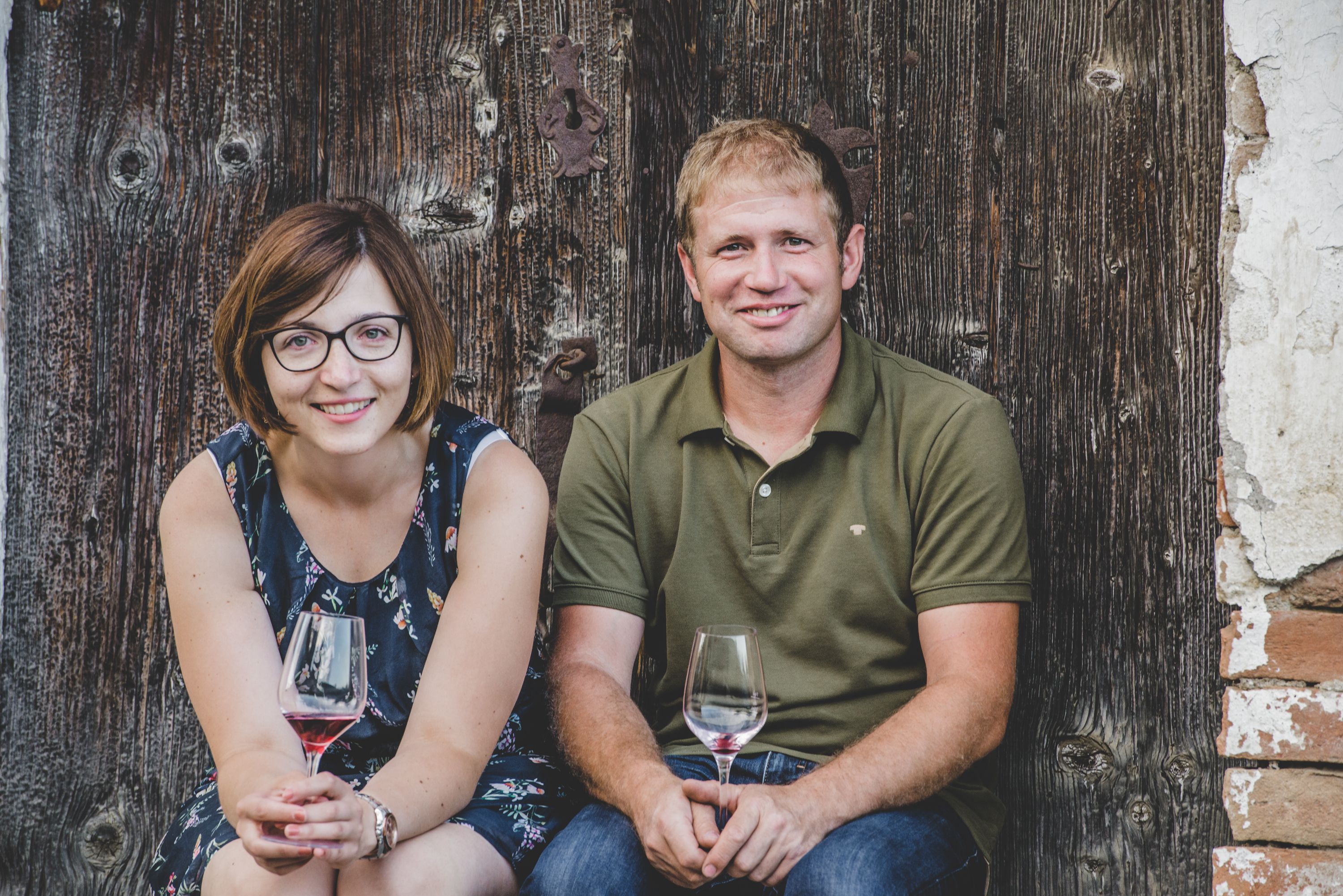 Muž a žena sedia pred drevenou stenou a v rukách držia poháre na víno.