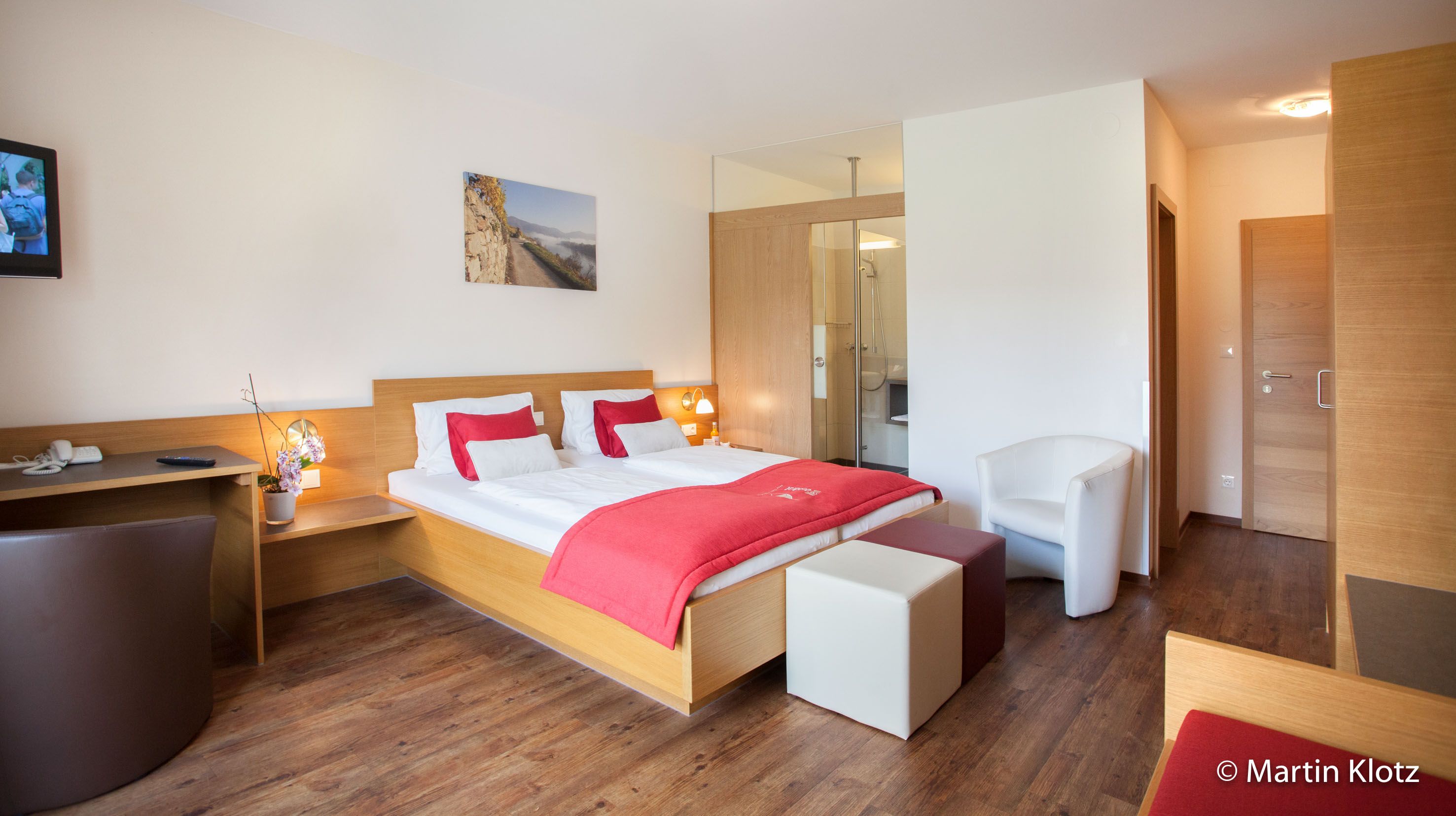 Moderná hotelová izba s manželskou posteľou, červenými akcentmi, písacím stolom a TV.