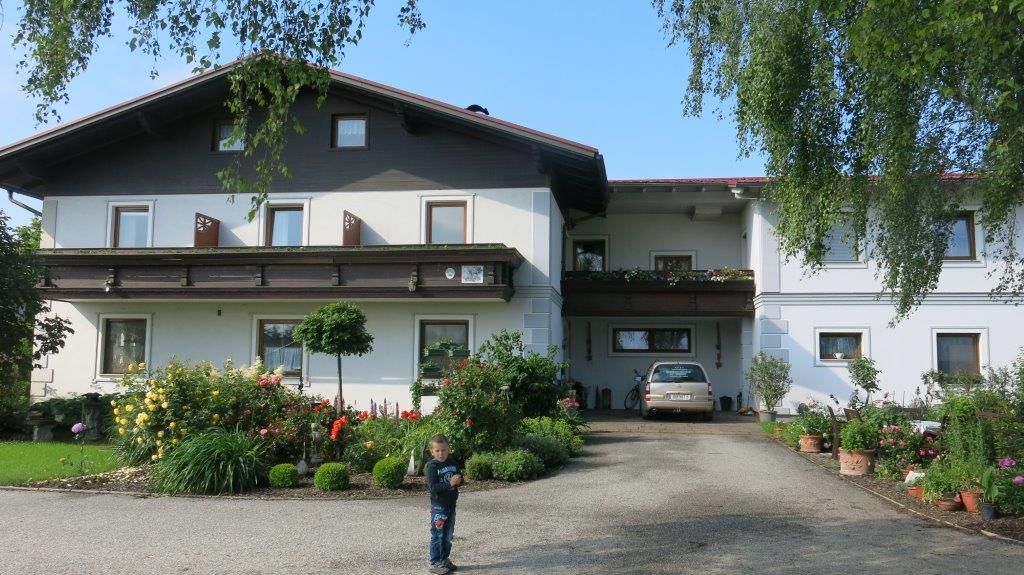Tradičný penzión s balkónom, obklopený udržiavanou záhradou. Na ceste pred domom stojí dieťa.