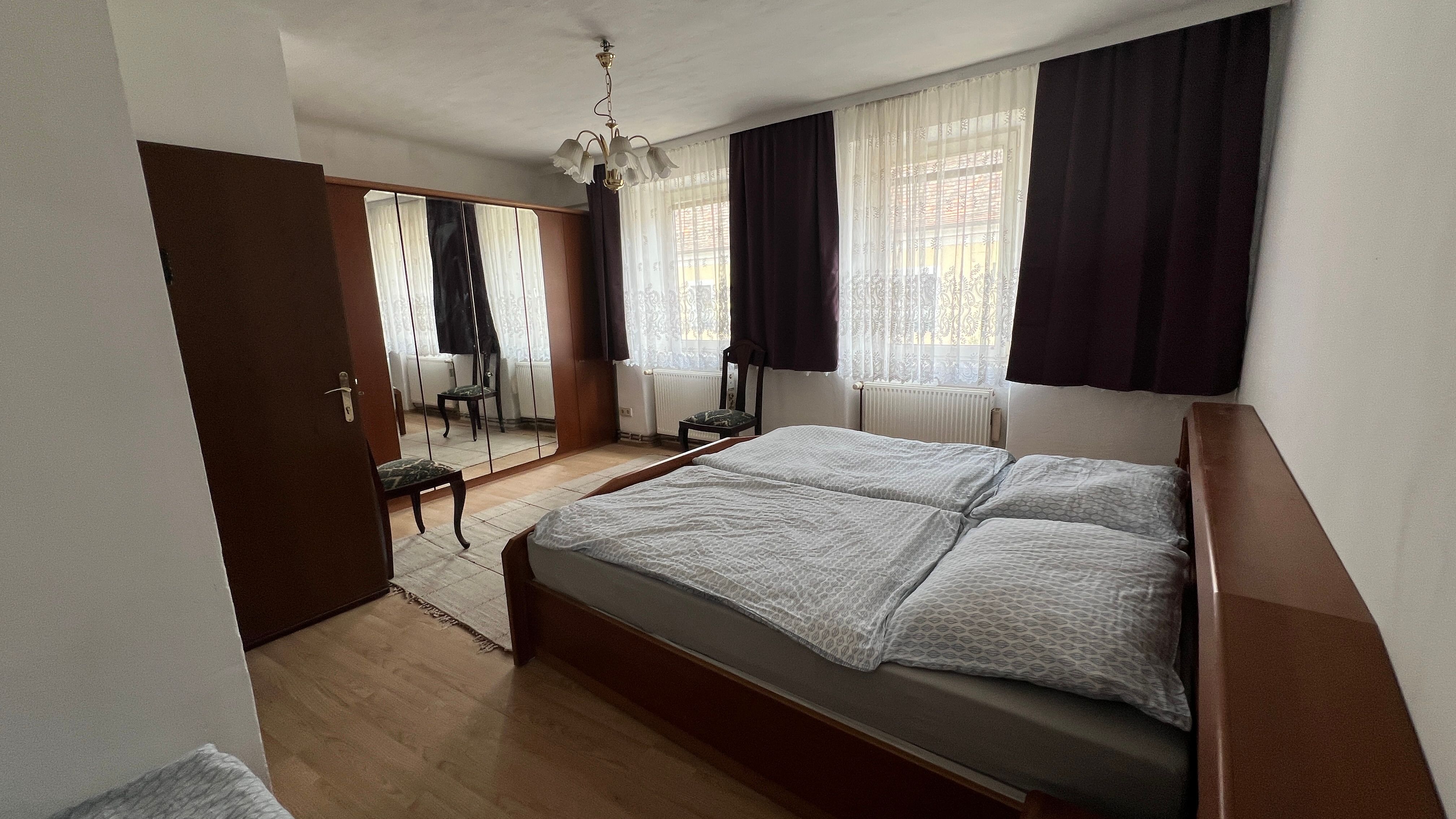 Spálňa s manželskou posteľou, šatníkovou skriňou so zrkadlom, stoličkou a závesmi na oknách.