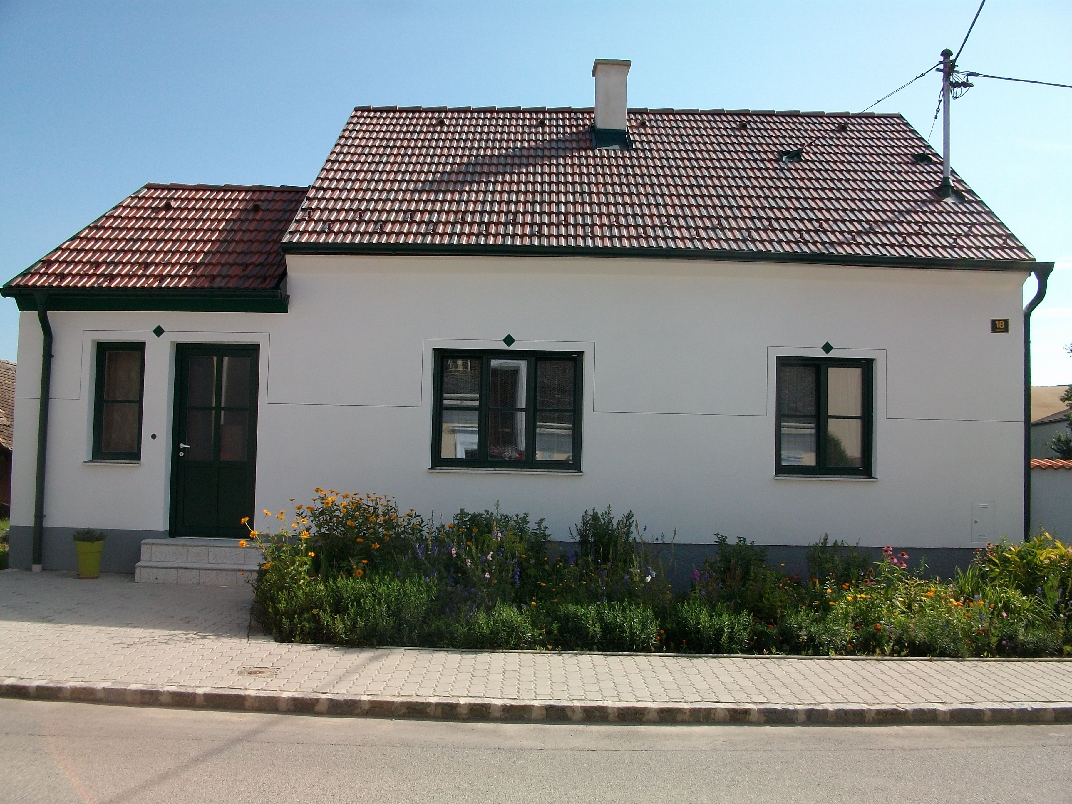 Malý biely domček s červenou škridlovou strechou a zeleným rámom dverí, obklopený udržiavanou záhradou s kvetmi.