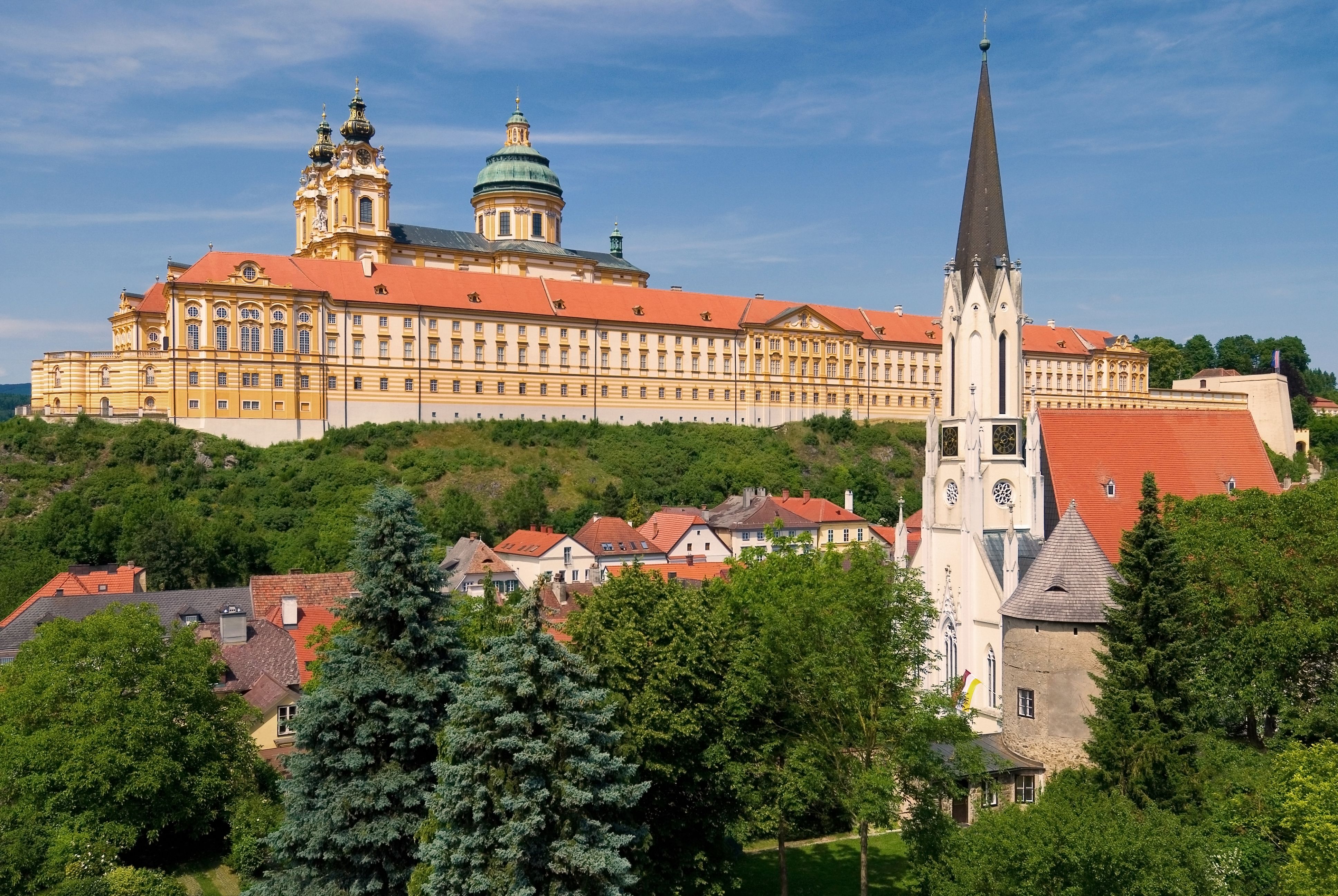 Pohľad na opátstvo Melk a farský kostol v Melku, obklopený zelenou krajinou a modrou oblohou.