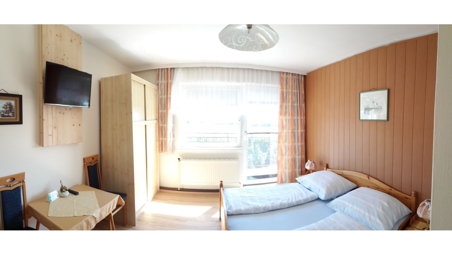 Útulná izba pre hostí s manželskou posteľou, dreveným nábytkom, malým stolíkom a TV na stene.