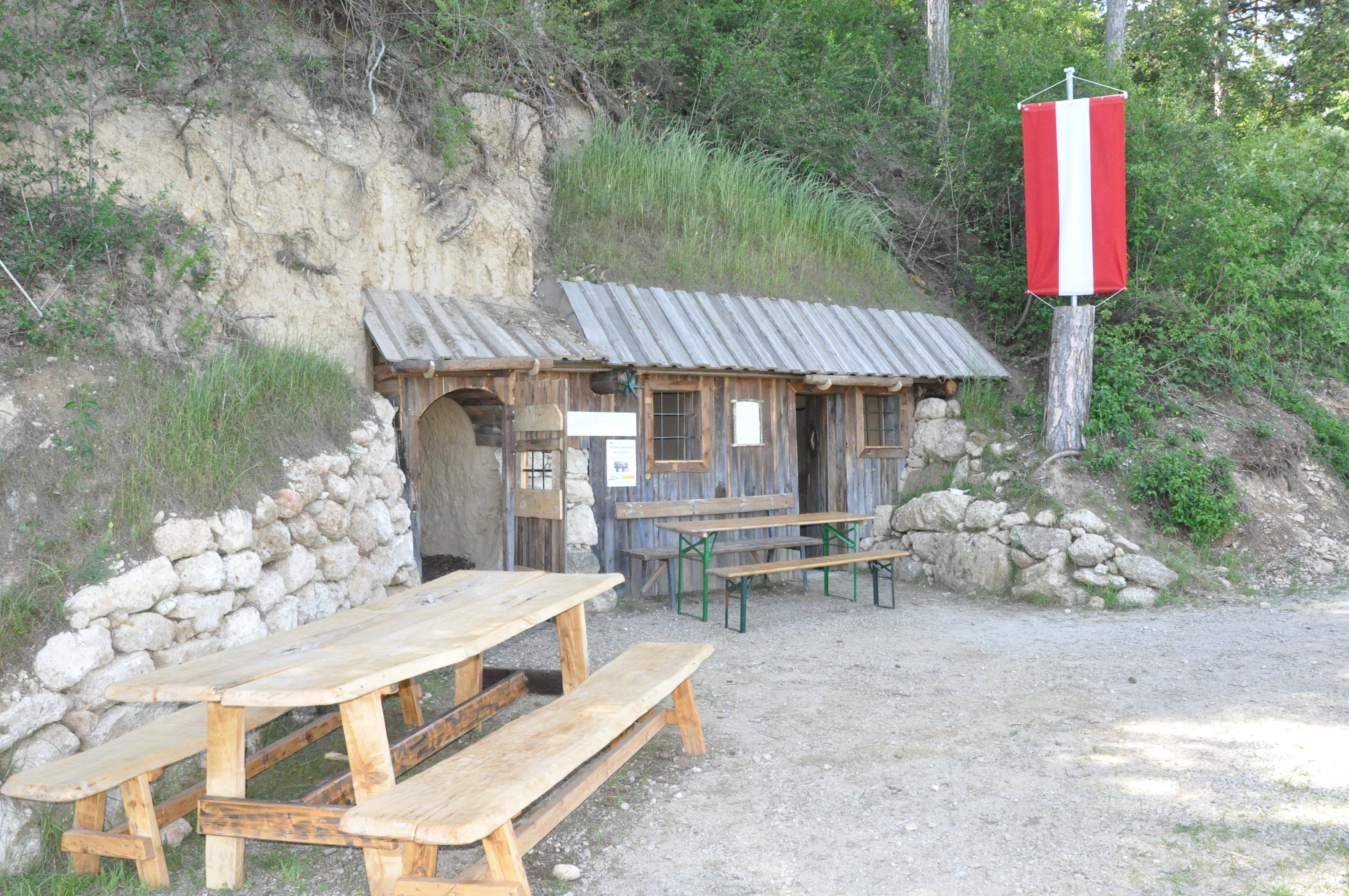 Rustikálna drevená chatka so stolom a lavicami pred ňou, postavená v kopci. Na strome visí červeno-biela vlajka.