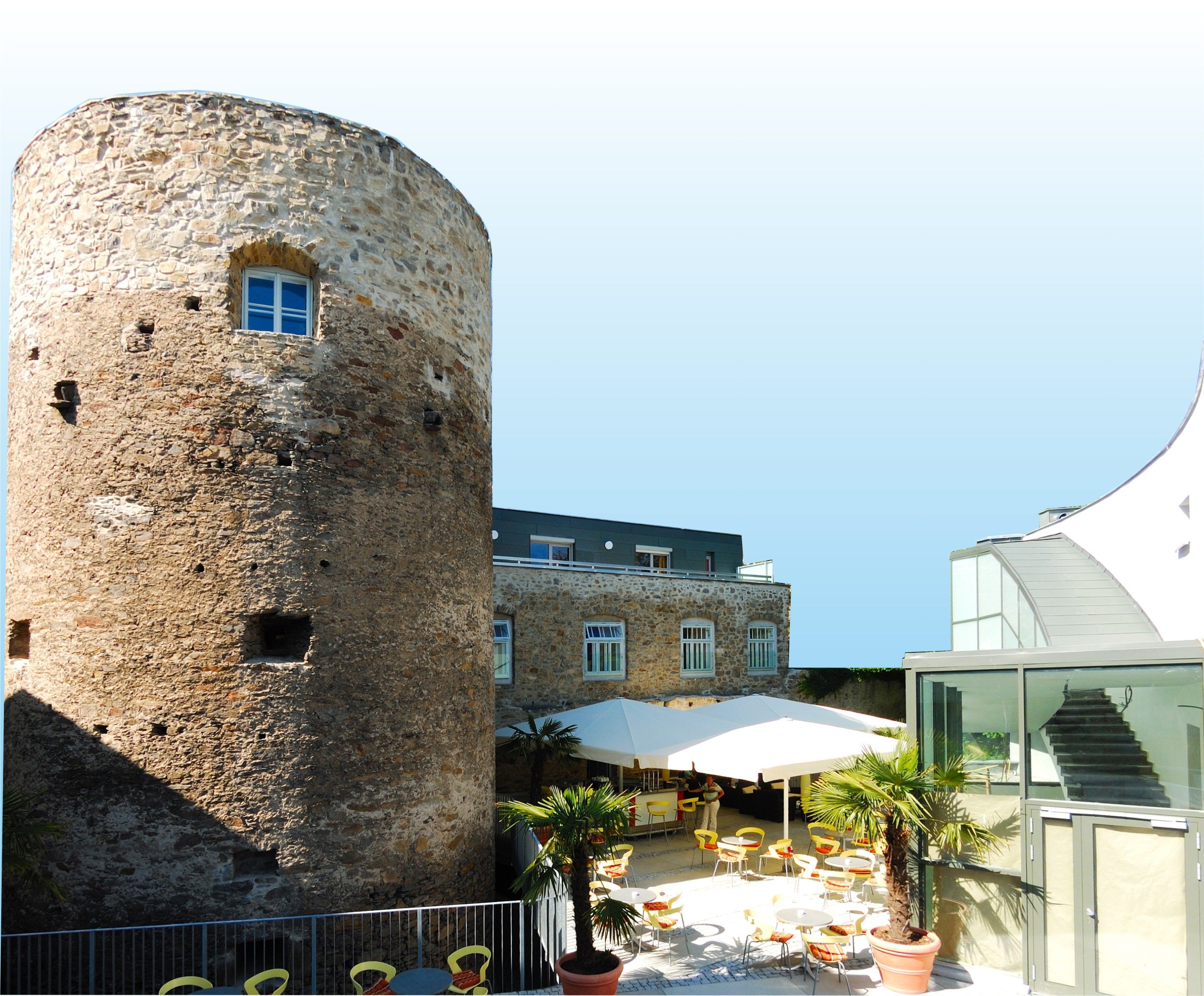 Historická veža vedľa modernej budovy s terasou a slnečníkmi.