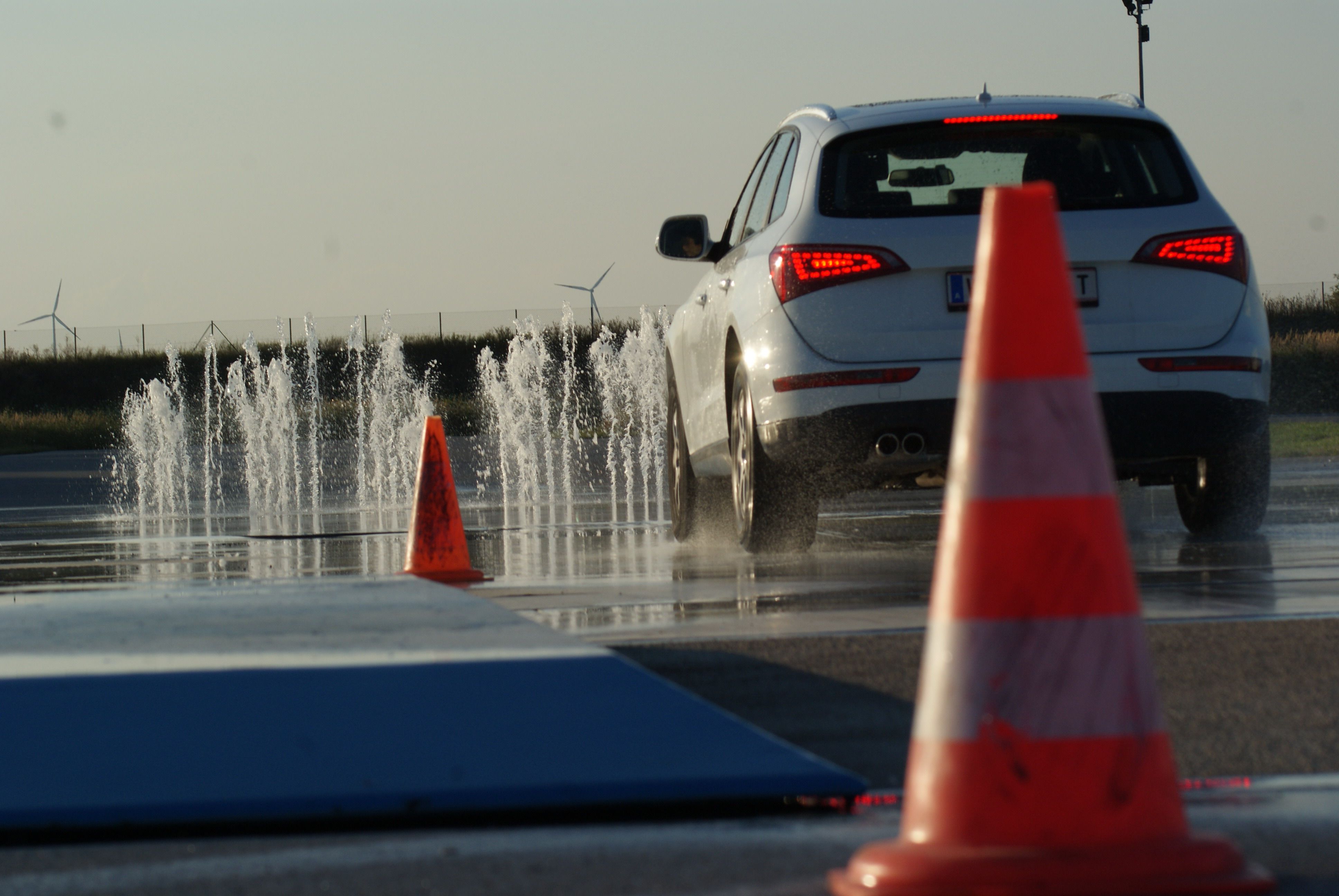 Auto jazdí po mokrej vozovke s vodnými fontánami a dopravnými kužeľmi.