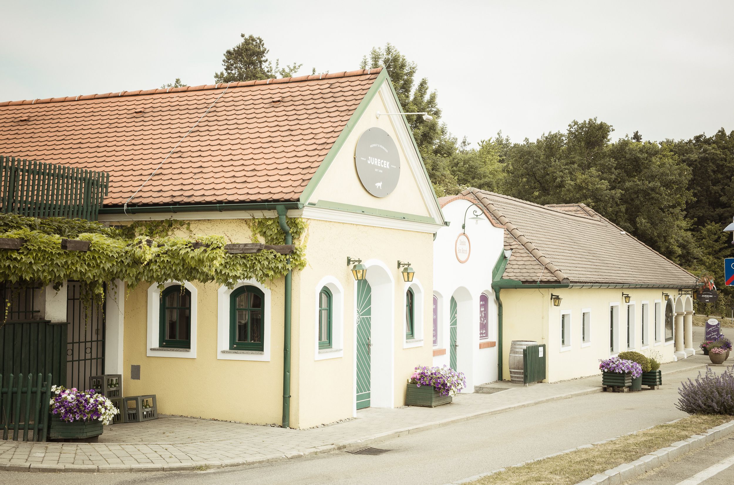 Exteriérový pohľad na Viechu so žltou fasádou a červenou strešnou krytinou, obklopenú rastlinami.