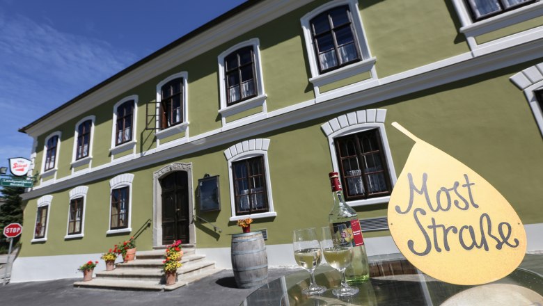 Zelen&aacute; budova s bielymi oknami, pred ňou st&ocirc;l s fľa&scaron;ou v&iacute;na a poh&aacute;rmi, tabuľka s n&aacute;pisom "Mu&scaron;t Stra&szlig;e".