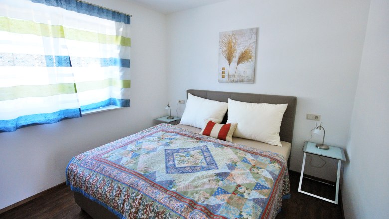 Sp&aacute;lňa s manželskou posteľou, farebn&yacute;m prehozom, dvoma nočn&yacute;mi stol&iacute;kmi a oknom s pruhovan&yacute;mi z&aacute;vesmi.