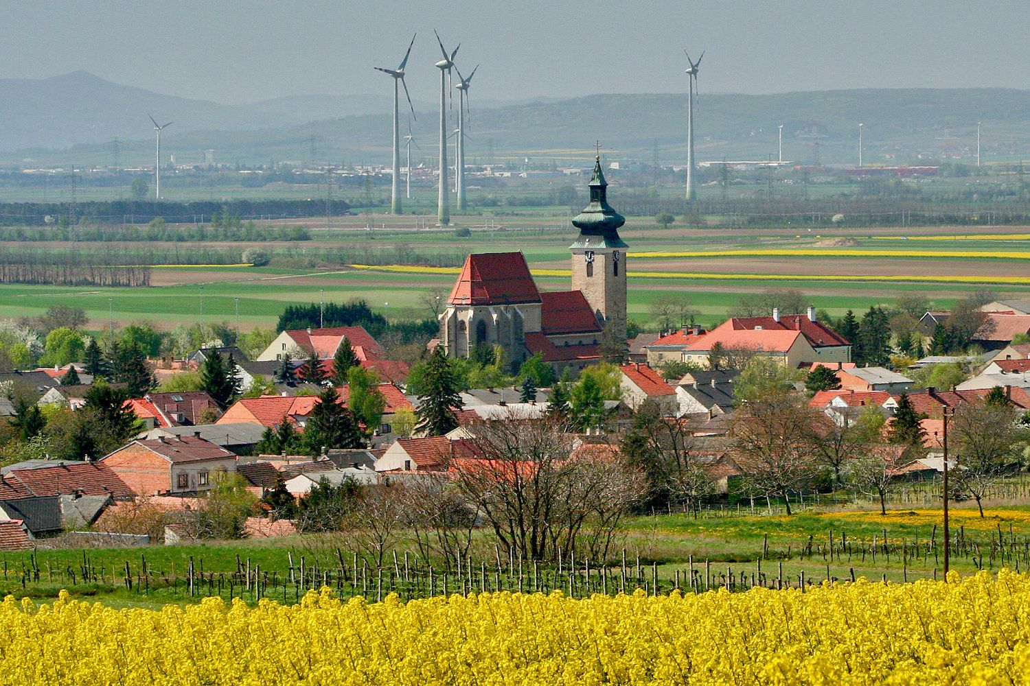 Pohľad na Pillichsdorf s kostolom, obklopený poľami a veternými turbínami v pozadí.