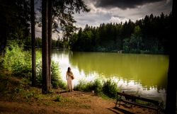 Ein ruhiger Moment am Ufer eines glitzernden Sees, umgeben von dichten Wäldern und sanften Hügeln. Die sanften Wellen spiegeln die dramatischen Wolken am Himmel wider und schaffen eine friedliche Atmosphäre, die zum Verweilen einlädt.