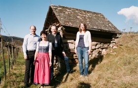 &Scaron;tyria ľudia stoja pred starou chatrčou na kopci s viničom v pozad&iacute;.