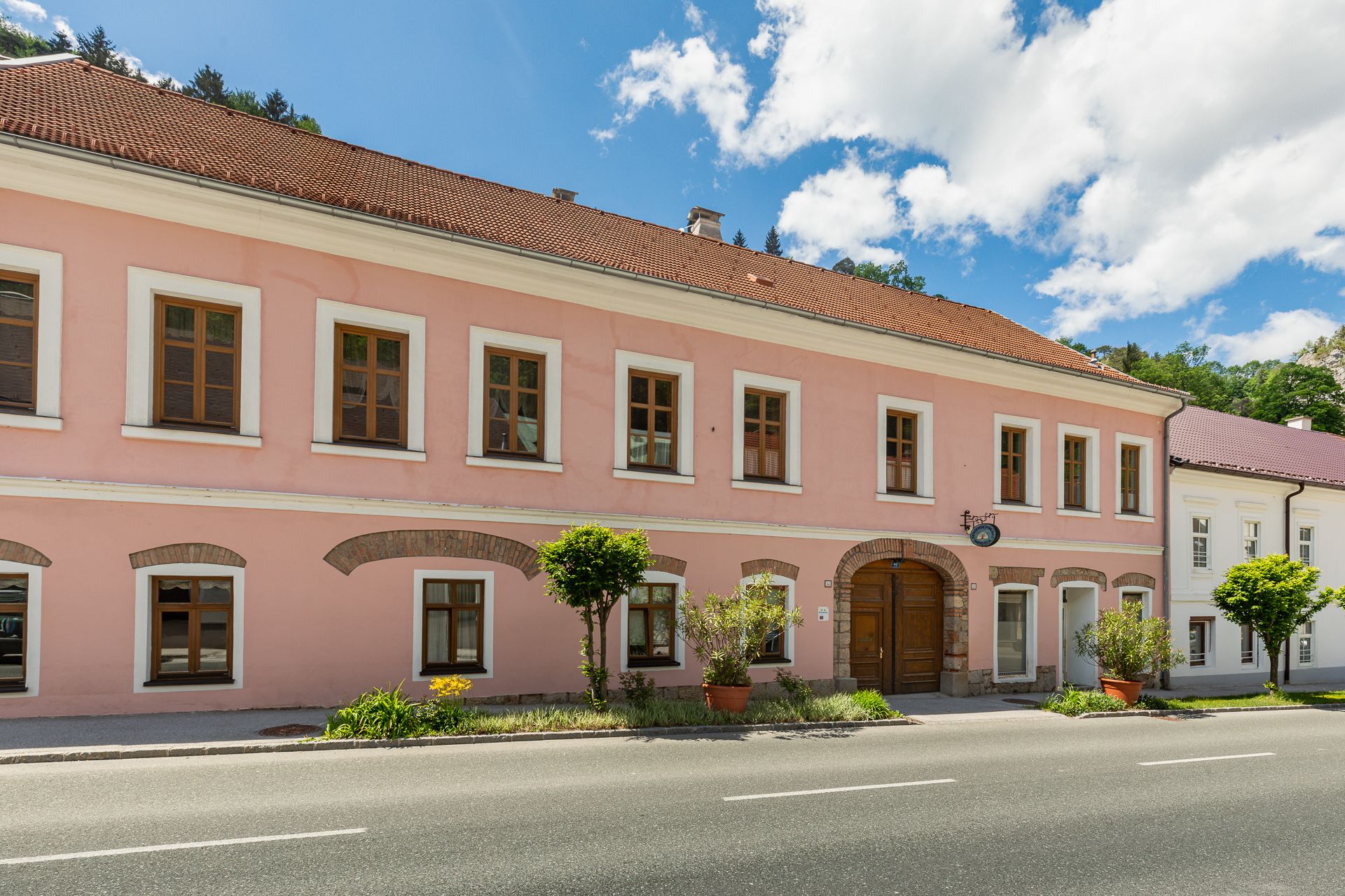 Dvojposchodová ružová budova s hnedými oknami a drevenými dverami, obklopená rastlinami, pod modrou oblohou.