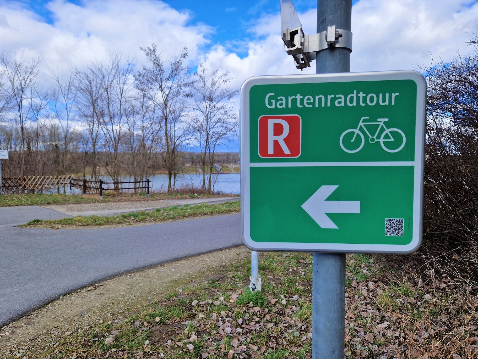 Zelená značka s nápisom "Gartenradtour" a symbolom bicykla ukazuje doľava. V pozadí je vidieť cestu a jazero.