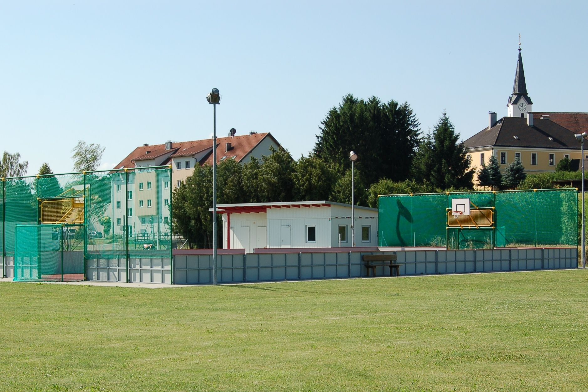 Zábavné ihrisko s basketbalovým košom a zelenou sieťou vedľa malej budovy. V pozadí vidieť obytné budovy a kostol.