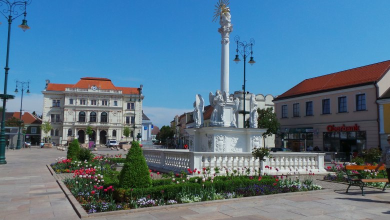 Hauptplatz mit barocker Säule und Blumenbeeten, im Hintergrund ein historisches Gebäude.