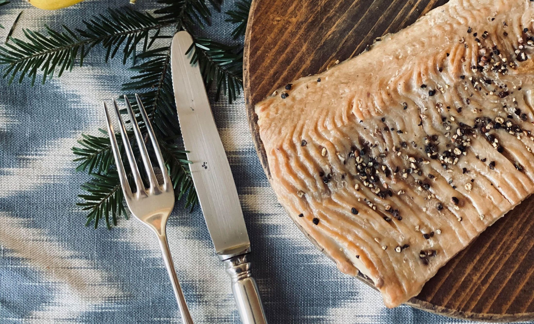 Detailný záber na údené filety z kapra na drevenej doske, zdobené zrnkami korenia. Vedľa neho sú na modrej utierke vidlička a nôž.