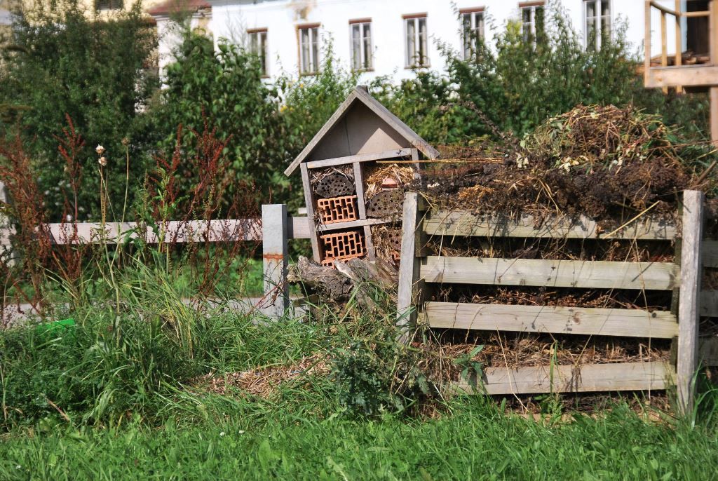 Hmyzí hotel vedľa kompostu v záhrade so zelenou trávou a stromami v pozadí.