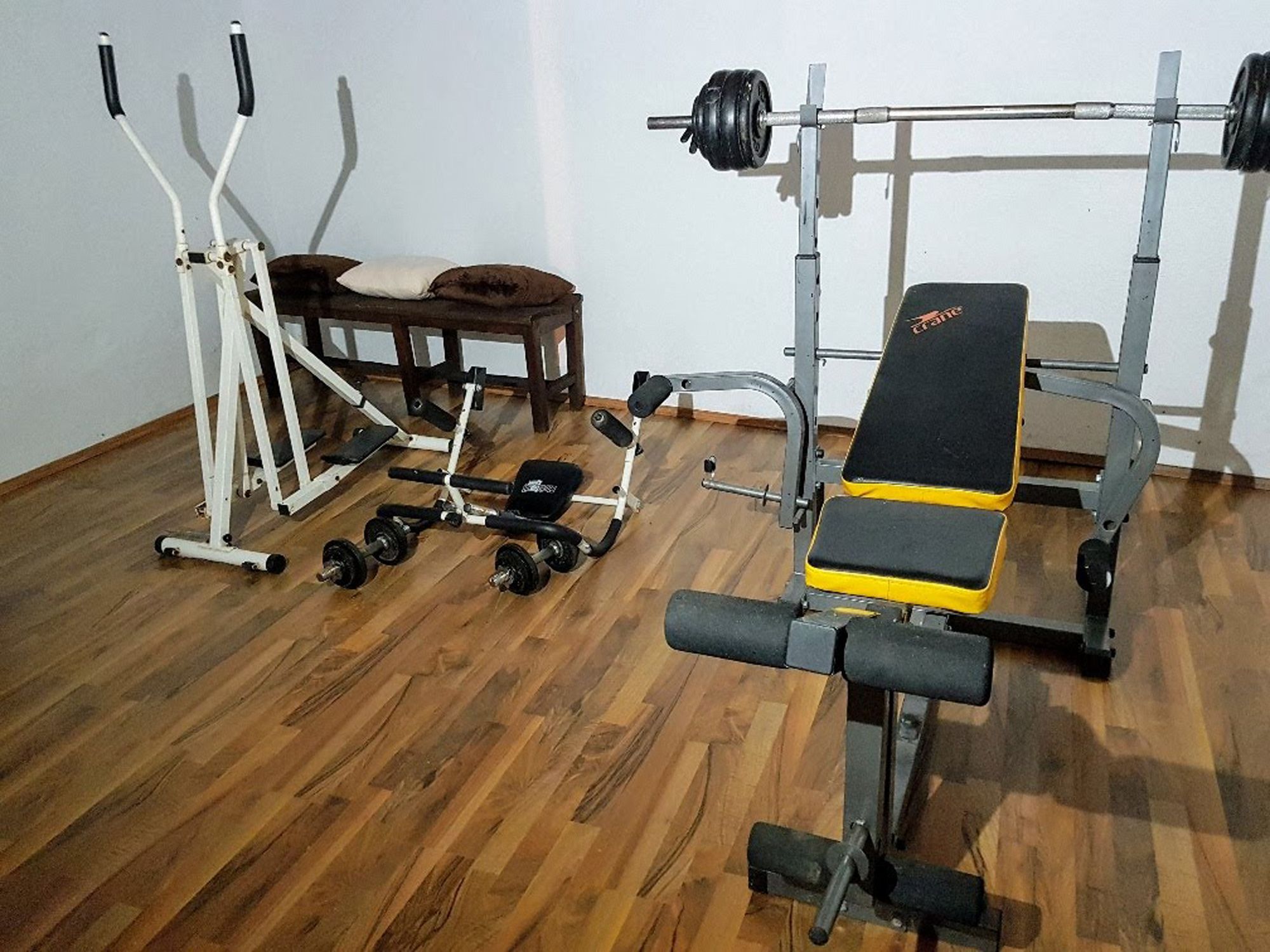 Posilňovňa s posilňovacou lavicou, crossovým trenažérom a činkami na drevenej podlahe.