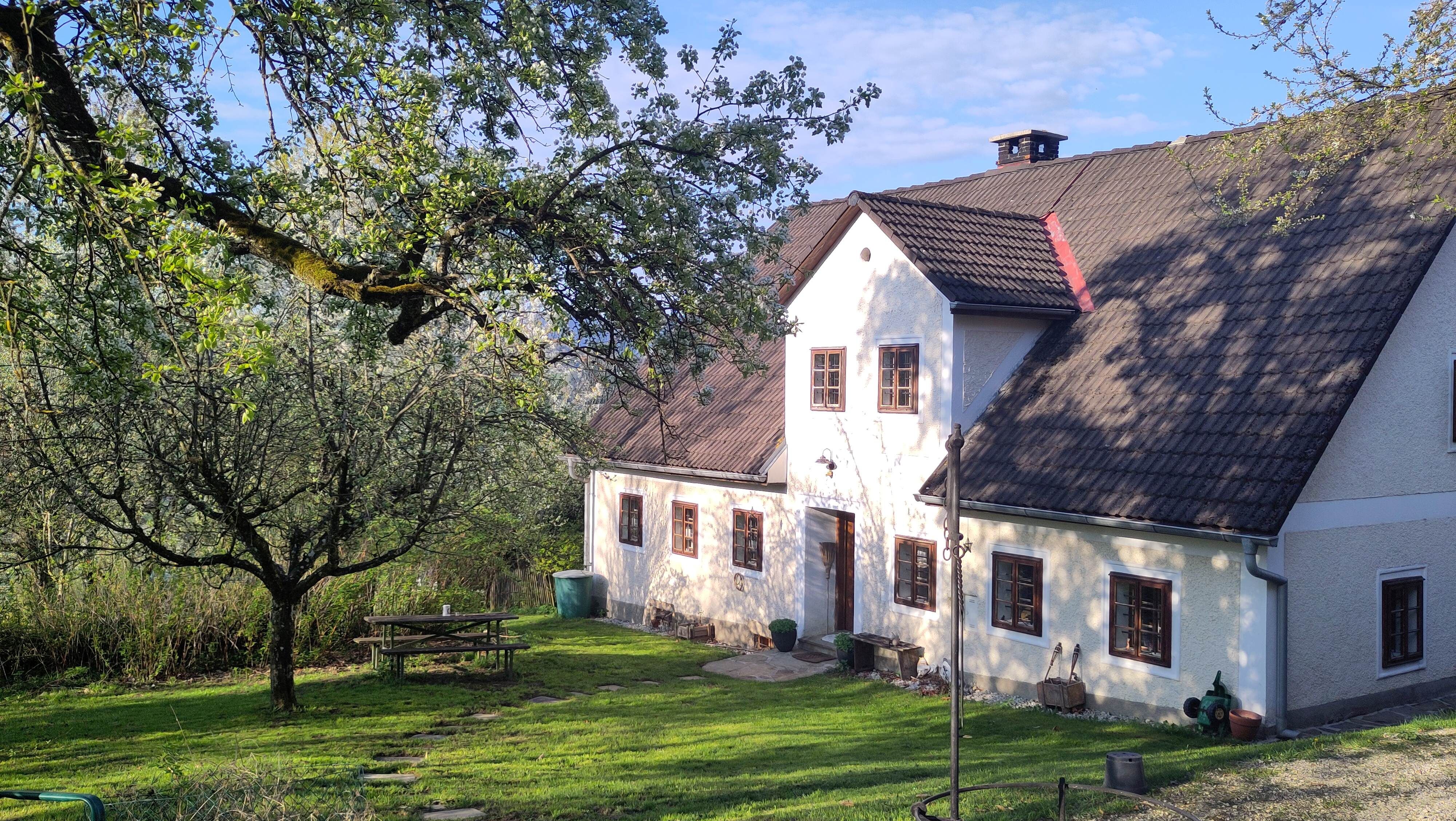 Tradičný vidiecky dom s bielymi stenami a hnedými oknami, obklopený zelenými stromami a upravenou záhradou s piknikovým stolom.