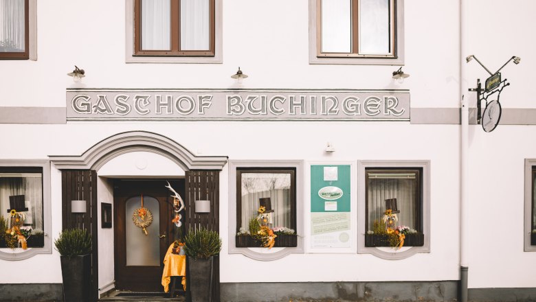 Fasáda hostinca s nápisom "Gasthof Buchinger". Vchod zdobia dekorácie a rastliny.