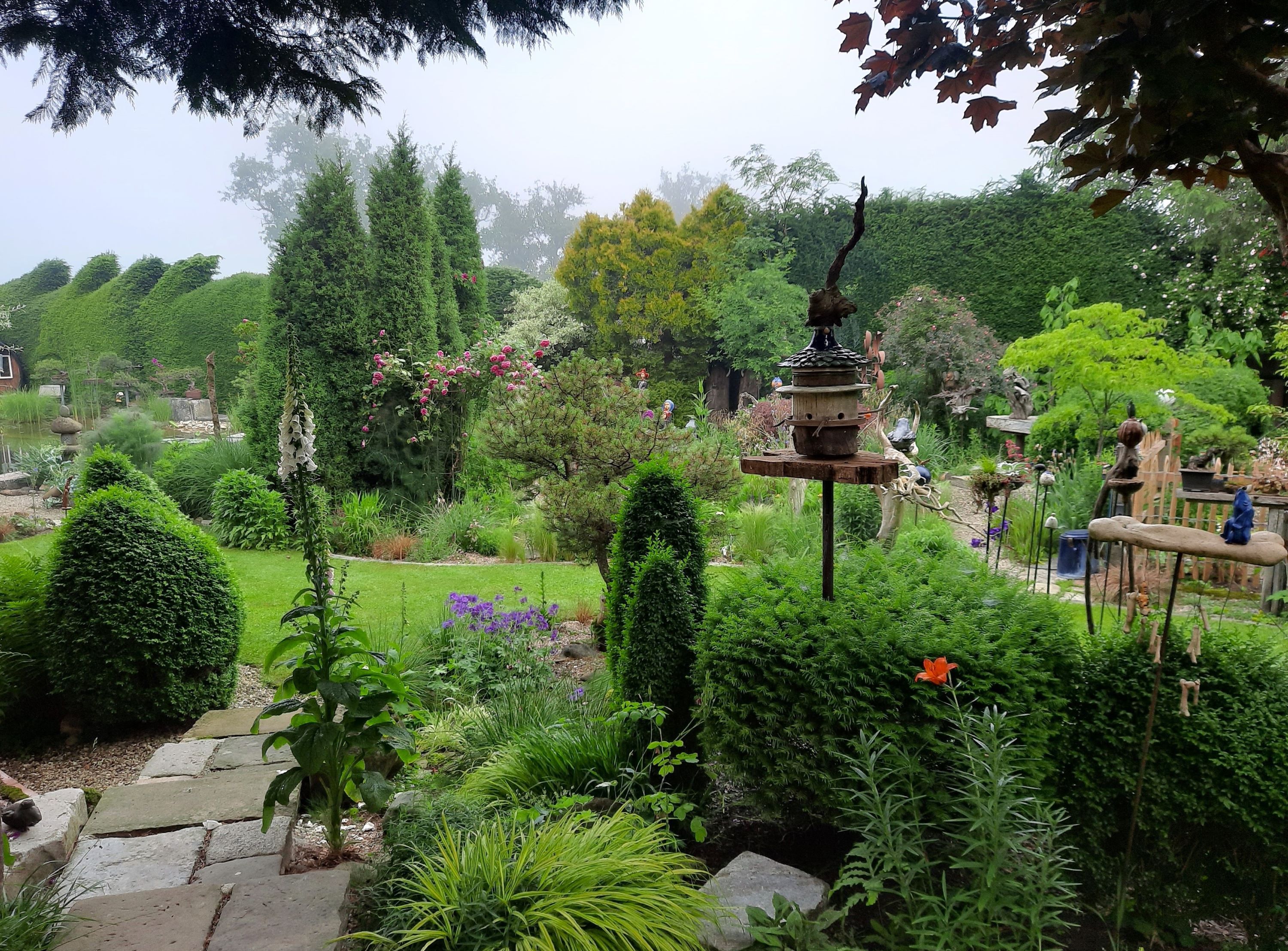 Bujná záhrada s rôznymi rastlinami, stromami a dekoráciami. V popredí stojí vtáčia búdka.