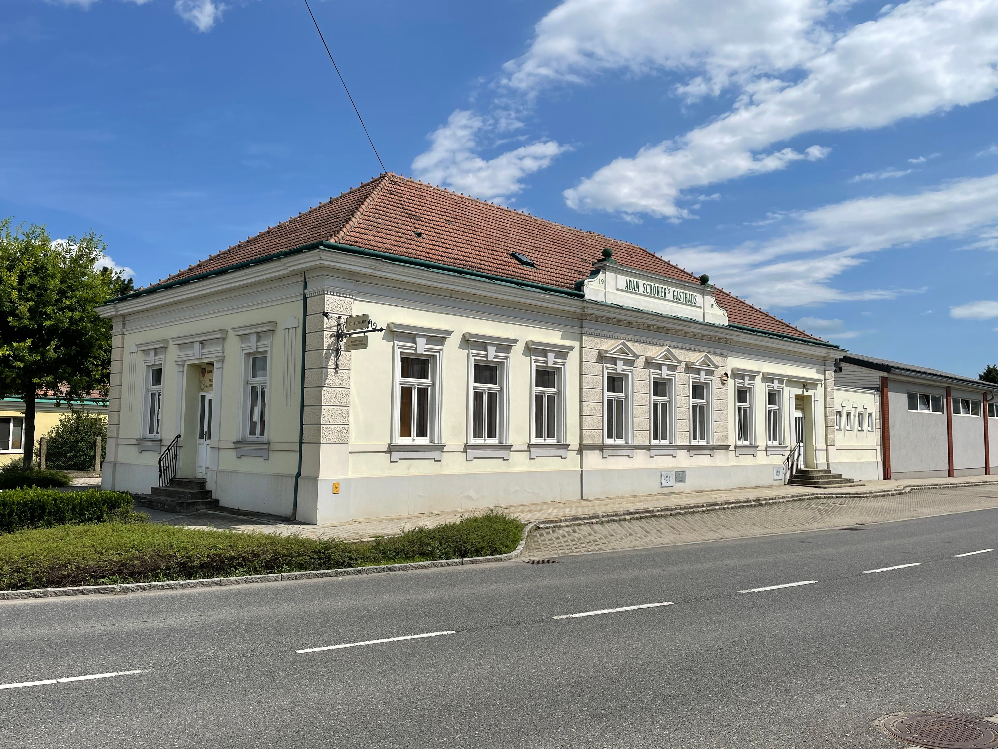 Kultúrny dom, svetlá historická budova s červenou tehlovou strechou, stojí pri ceste pod modrou oblohou.