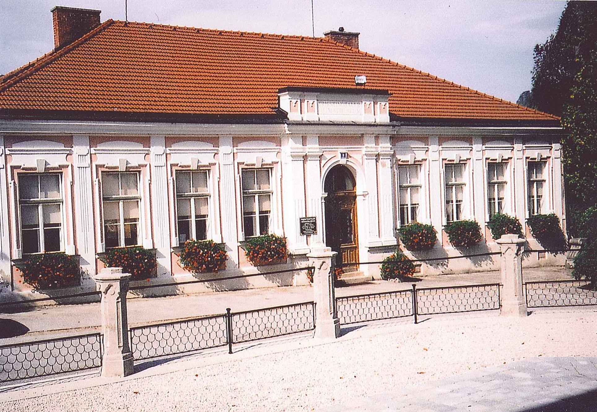 Historická budova s červenou škridlovou strechou a bielymi okennými rámami, obklopená kvetmi.