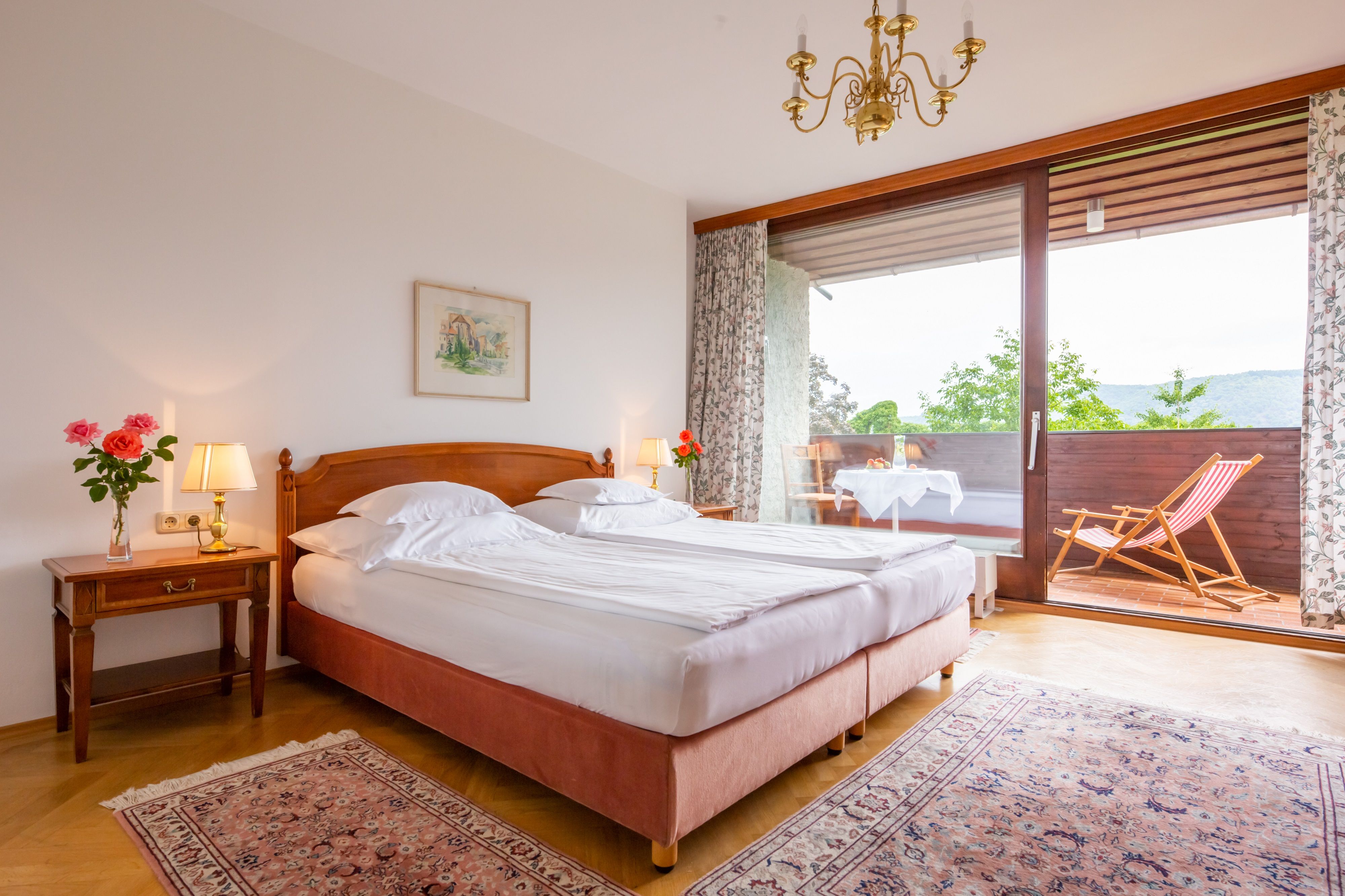 Útulná spálňa s manželskou posteľou, nočnými lampami a balkónom s ležadlom a stolom.