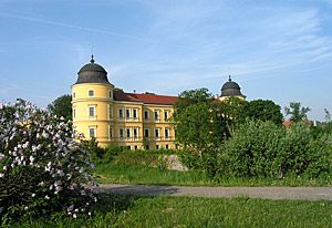 Žltý hrad s dvoma vežami a červenou strechou, obklopený stromami a kvetmi.