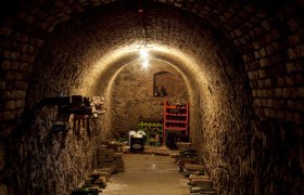 &Uacute;zka klenut&aacute; v&iacute;nna pivnica s tehlov&yacute;mi stenami a uložen&yacute;mi fľa&scaron;ami.