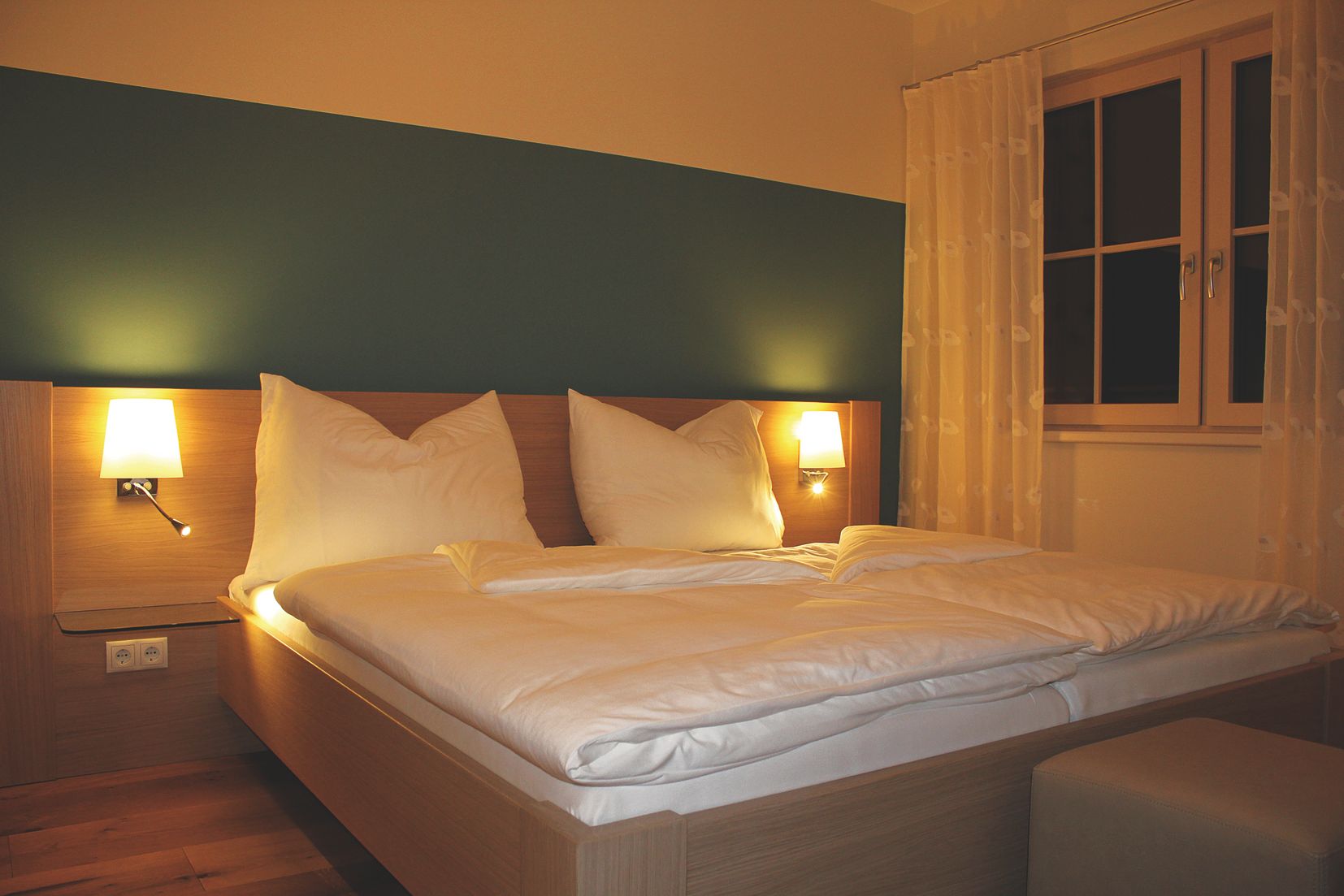 Útulná izba pre hostí s manželskou posteľou, dvoma nočnými lampami a oknom so závesmi.