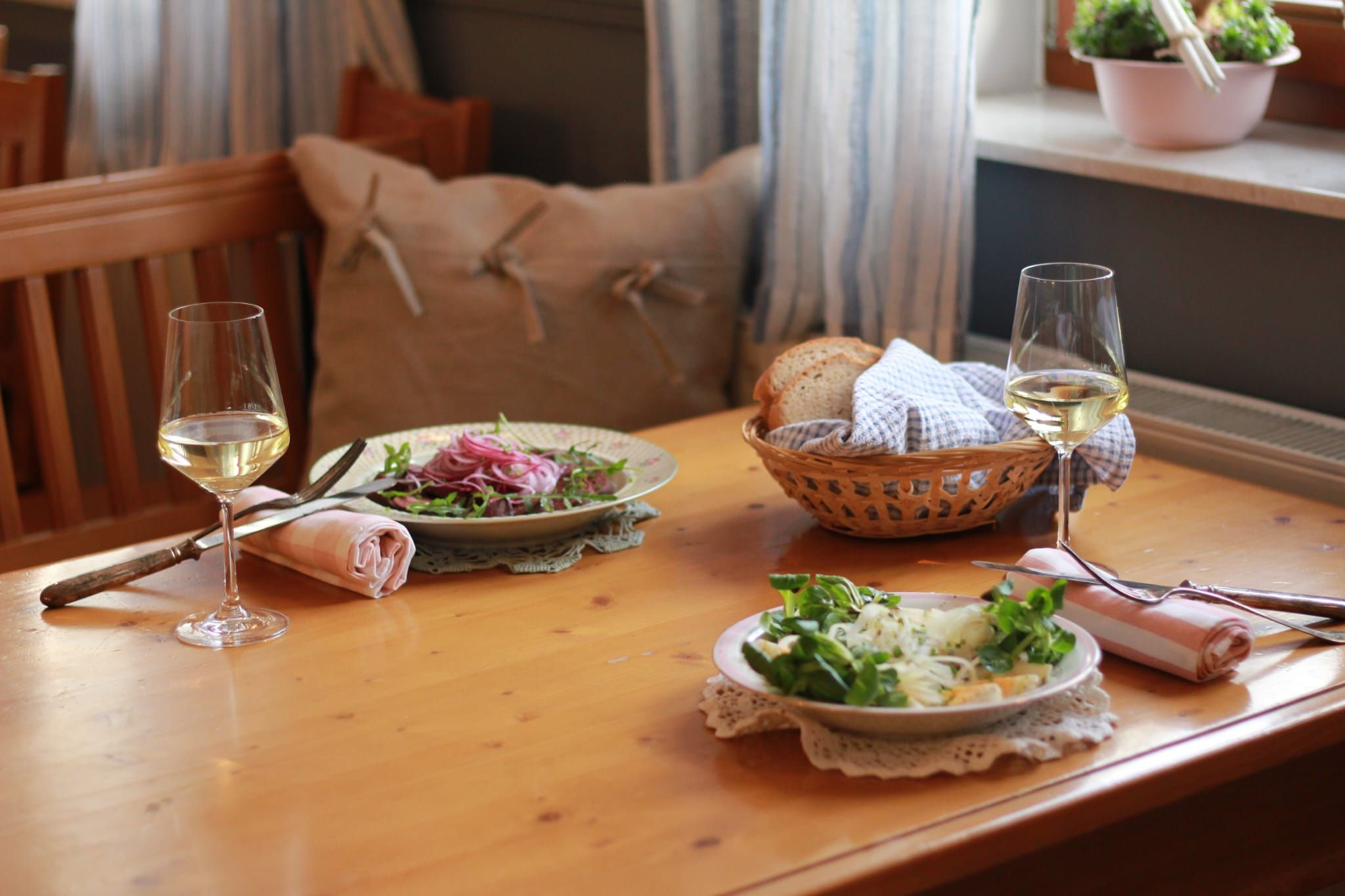 Dva taniere šalátu a dva poháre bieleho vína na drevenom stole, vedľa nich košík s chlebom.