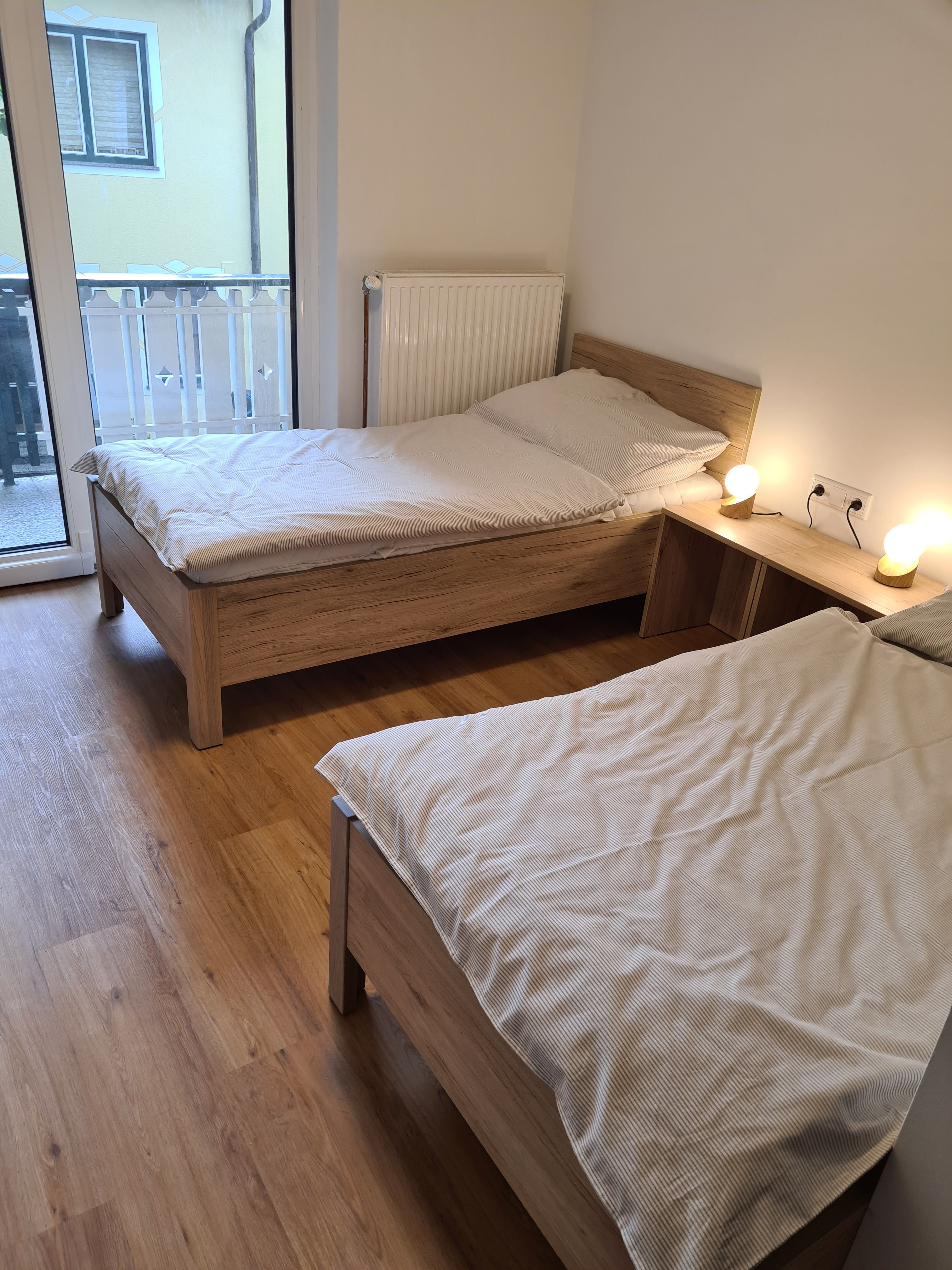 Dve samostatné postele v svetlej izbe s drevenou podlahou a prístupom na balkón.