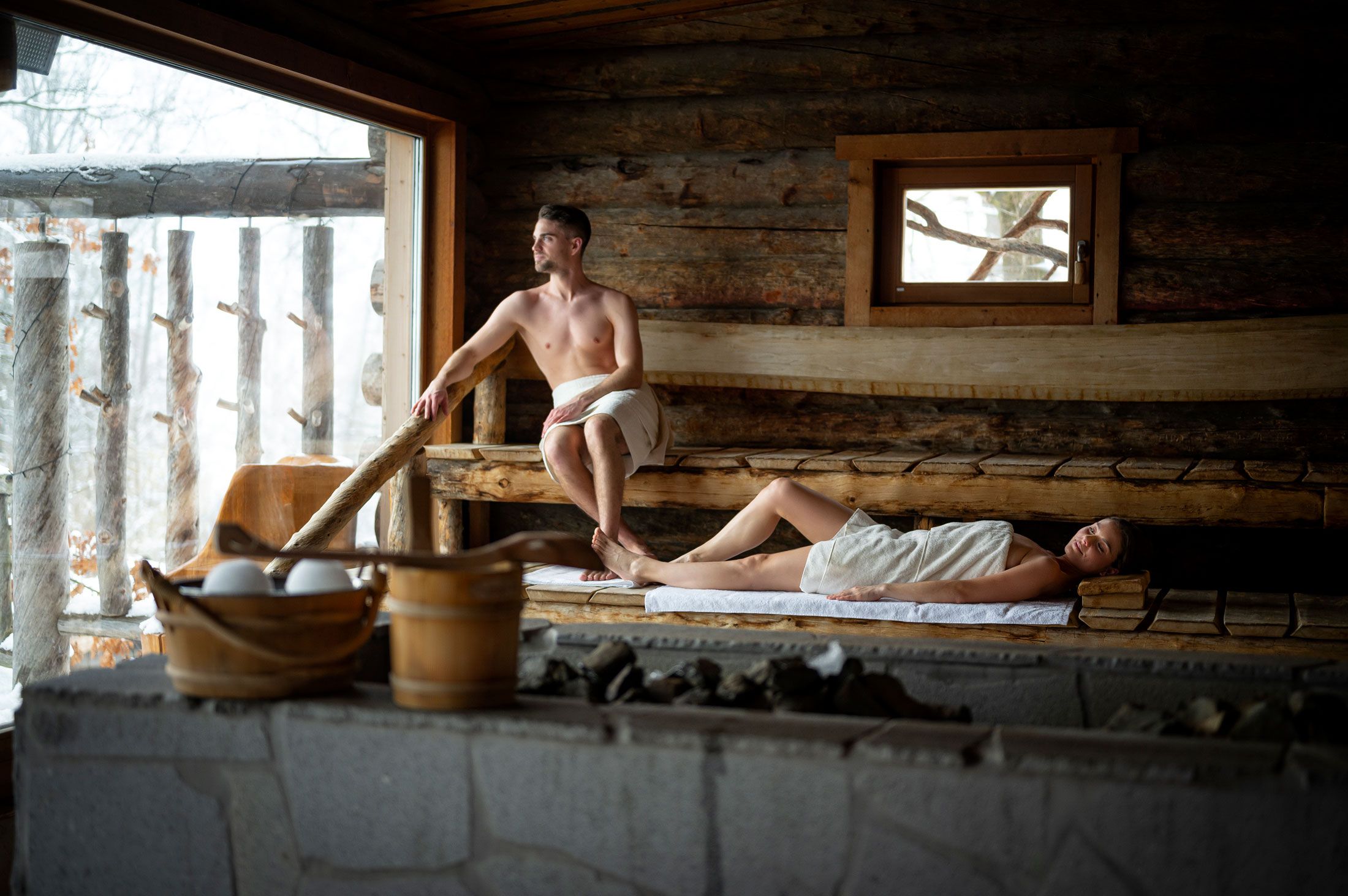Dvaja ľudia v drevenej saune, jeden sedí, druhý leží, obaja sú zabalení v uterákoch.