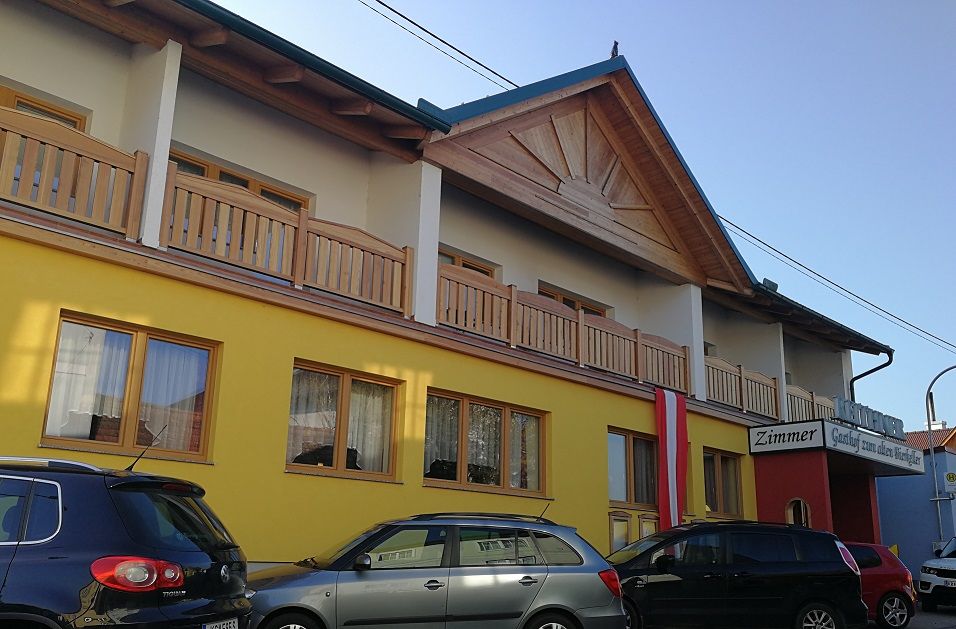 Pohľad zvonku na žltú budovu s drevenou verandou a autami pred ňou.