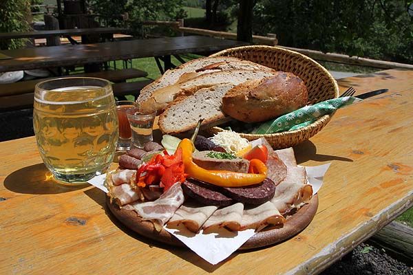 Občerstvenie s chlebom, klobásou, slaninou a nápojmi na drevenom stole vonku.